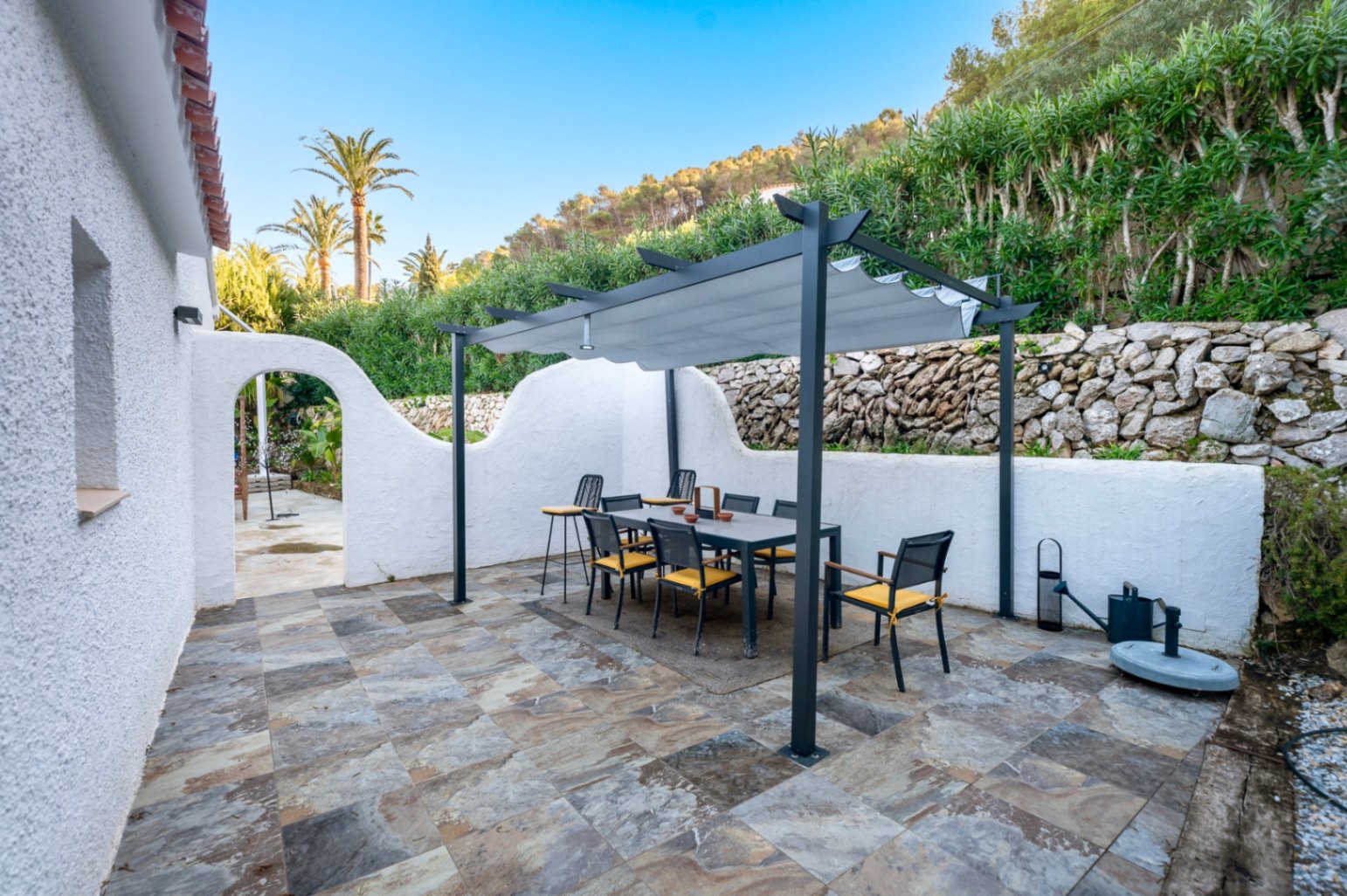 Espace repas extérieur avec pergola moderne, dallage en pierre naturelle et murs blanchis à la chaux, entouré d'une végétation méditerranéenne.