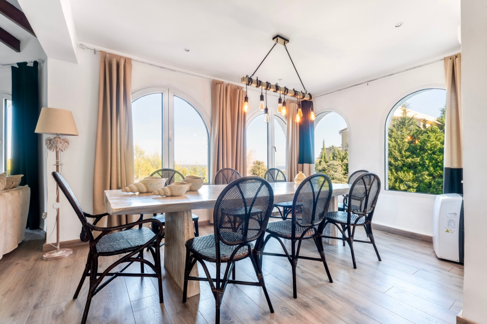 Salle à manger lumineuse avec table en bois massif, chaises en rotin noir et fenêtres cintrées. Un luminaire suspendu de style industriel complète l'espace.