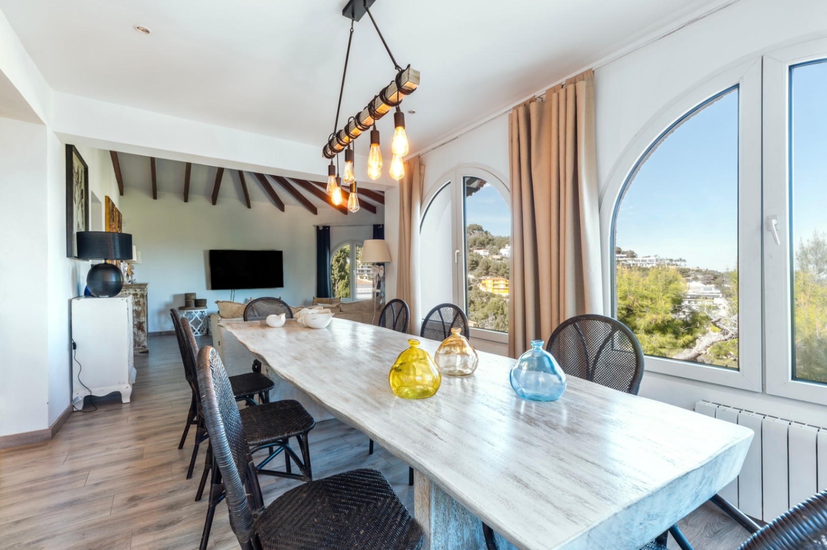 Salle à manger lumineuse avec table en bois rustique, chaises en osier noir et fenêtres en arc. L'espace dispose d'un luminaire industriel et d'un plafond à poutres apparentes.