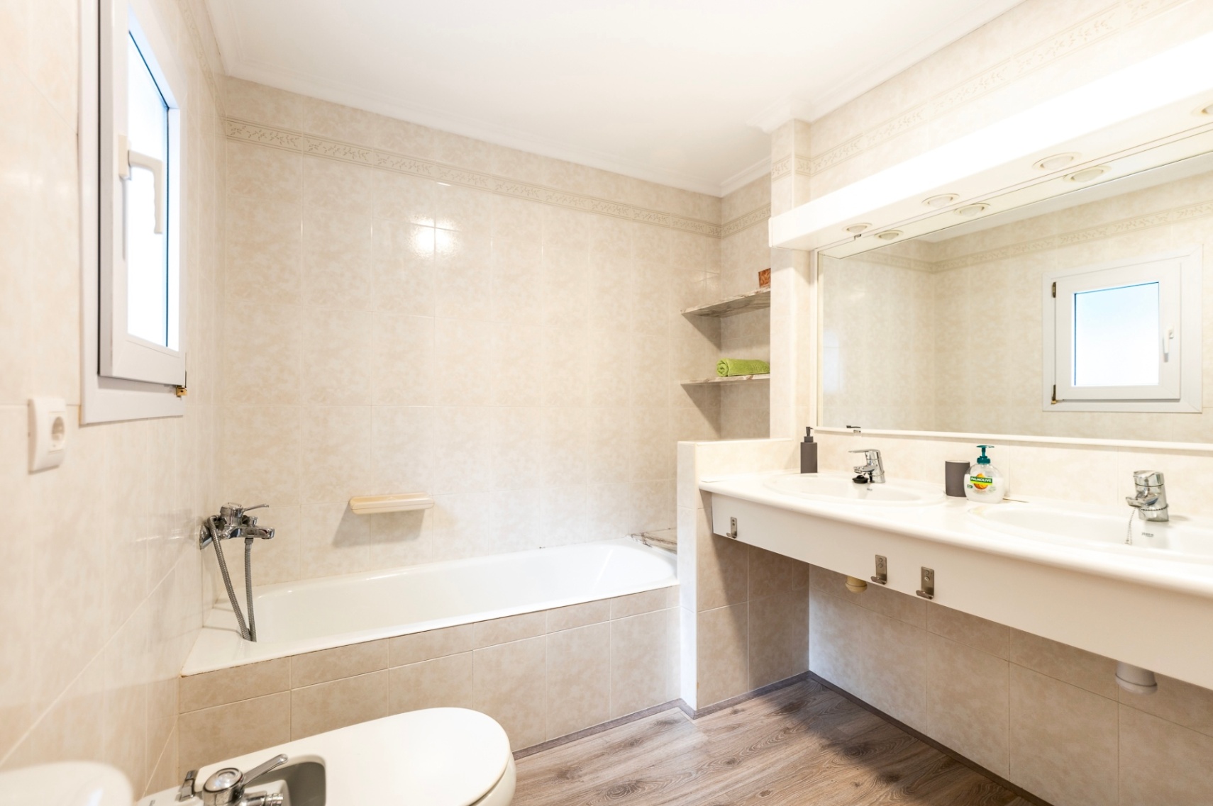Salle de bain avec baignoire encastrée, double vasque et sol imitation bois. Elle dispose de carrelage beige, d'un grand miroir éclairé et d'une fenêtre apportant de la lumière naturelle.