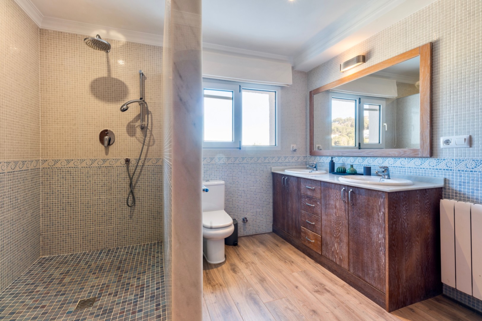 Salle de bain avec douche à l'italienne en mosaïque, double vasque sur meuble en bois et sol imitation parquet. Une fenêtre apporte une luminosité naturelle.