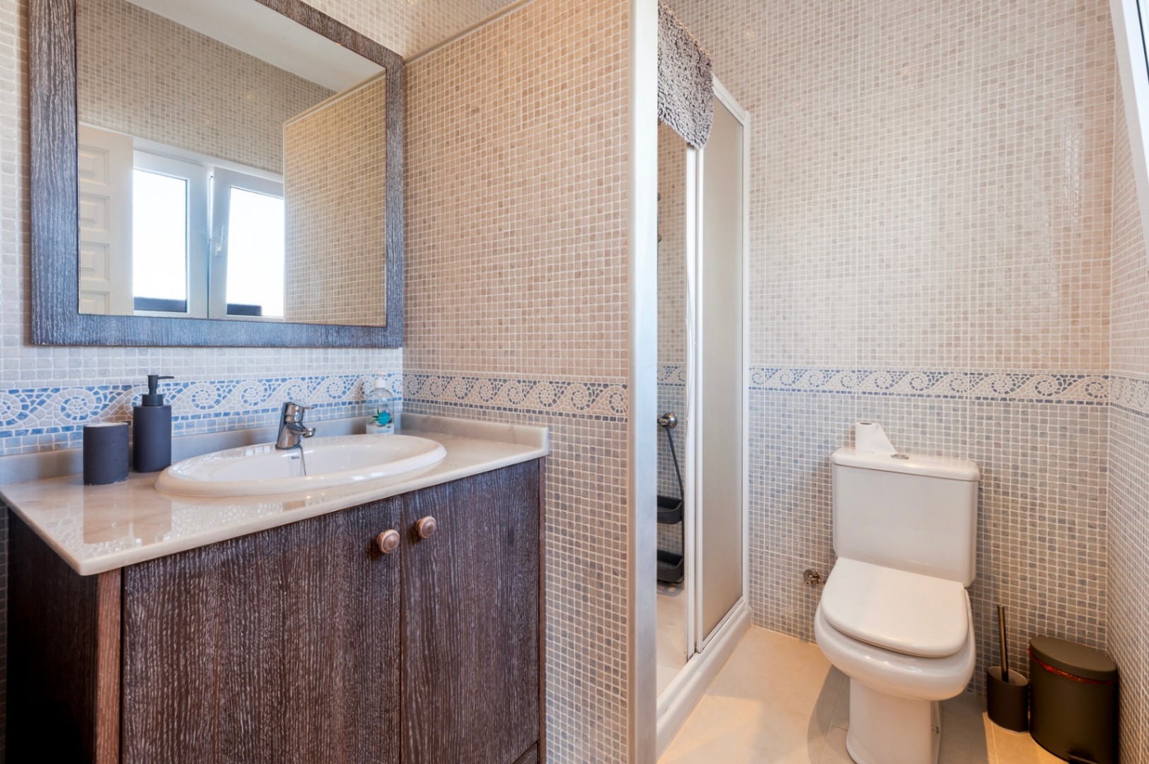 Salle de bain avec murs en mosaïque beige et frise bleue. Elle dispose d'un meuble vasque en bois sombre, d'un plan de travail en marbre et d'une douche vitrée.