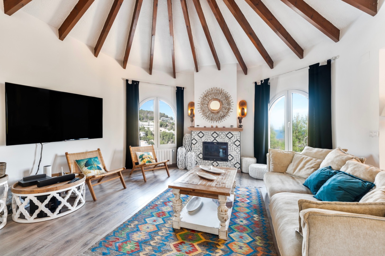 Salon circulaire avec plafond à poutres apparentes en éventail, cheminée ornée de carreaux à motifs et fenêtres cintrées ouvrant sur l'extérieur de la villa.