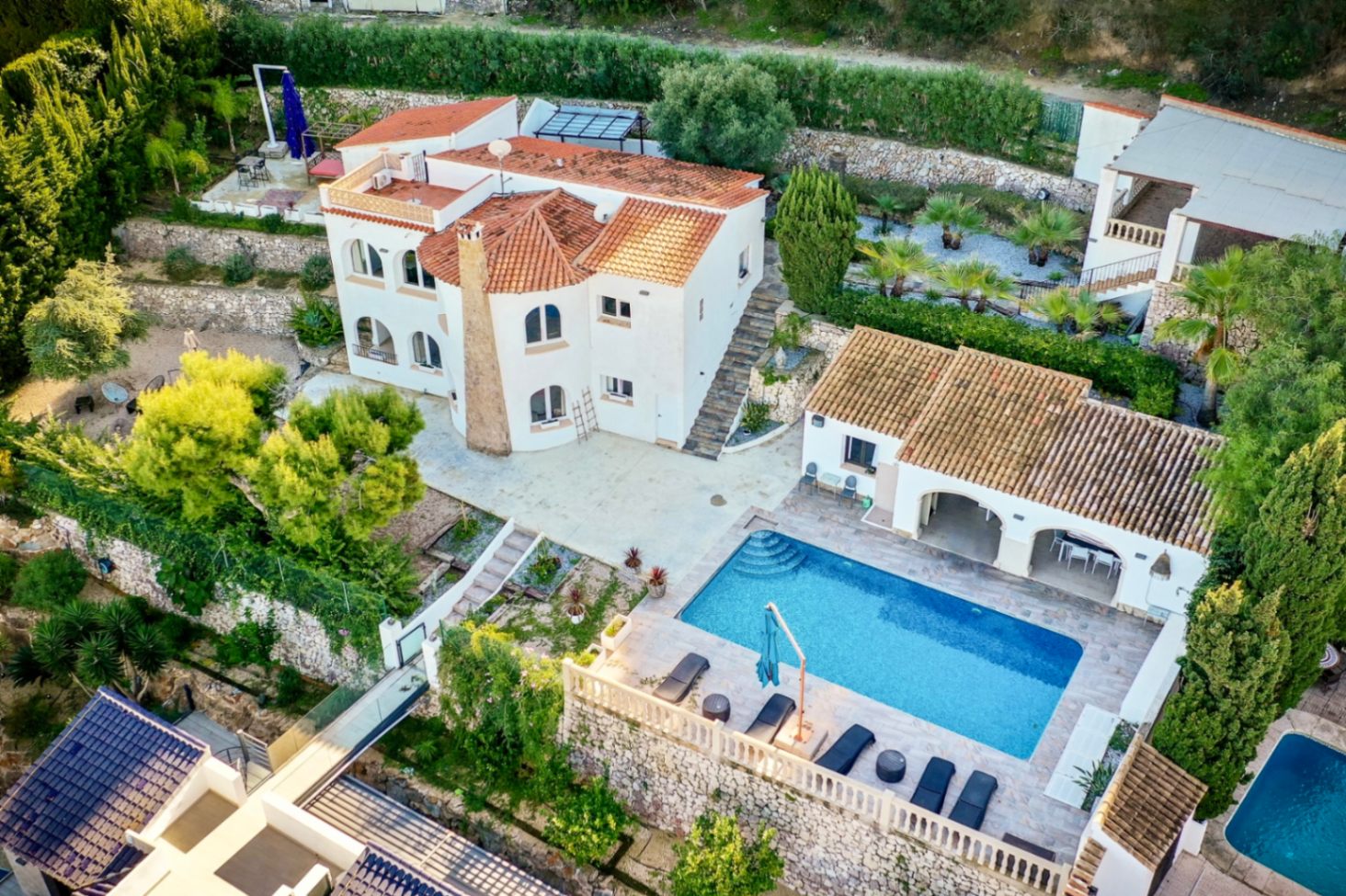 Vue aérienne de la villa aux façades blanches et toits en tuiles, avec sa piscine rectangulaire, sa terrasse en pierre et ses jardins méditerranéens en terrasses.