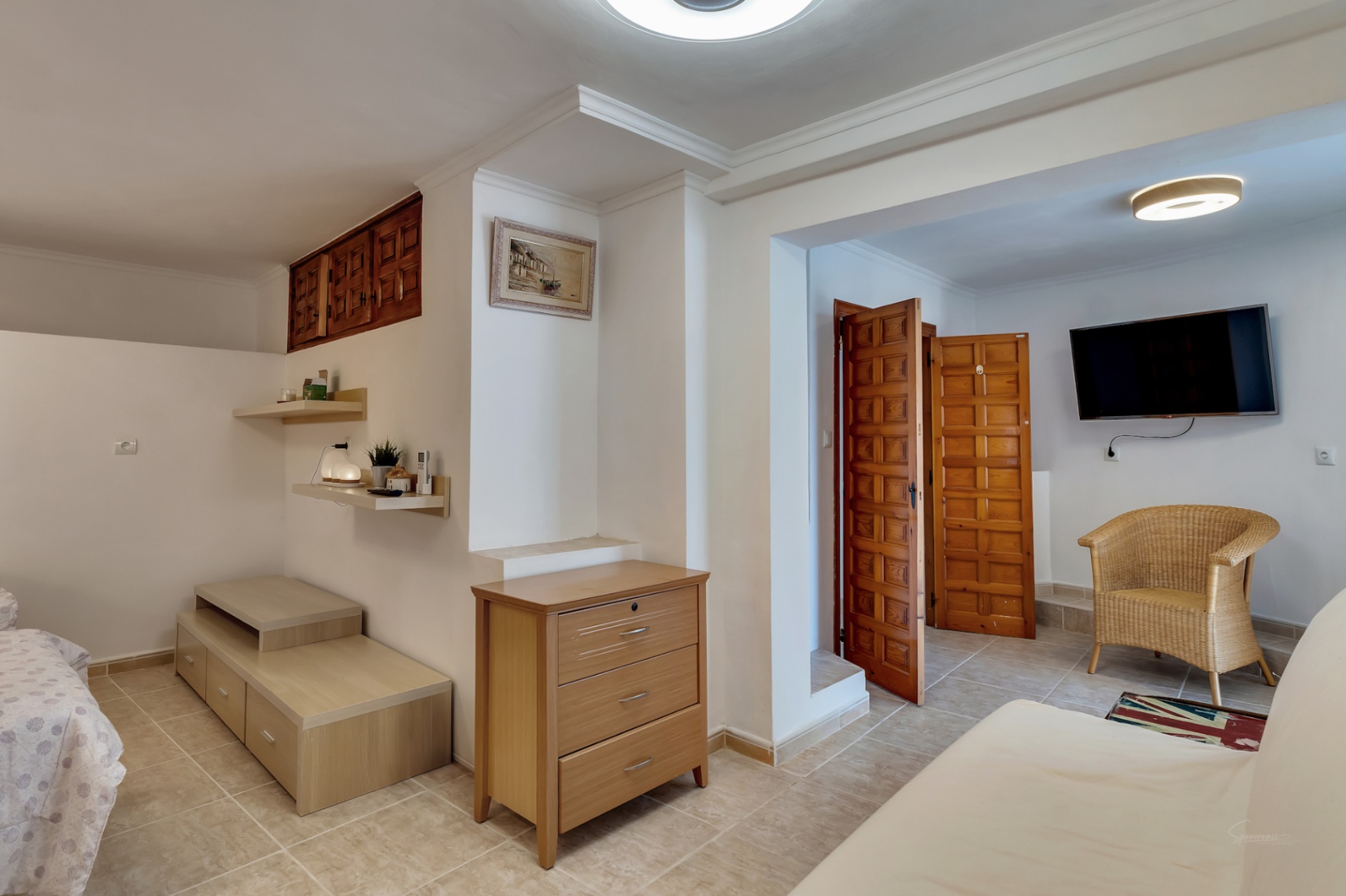 Chambre au niveau inférieur avec sol en grès cérame, portes en bois de style castillan, coin salon avec fauteuil en osier et rangements intégrés.