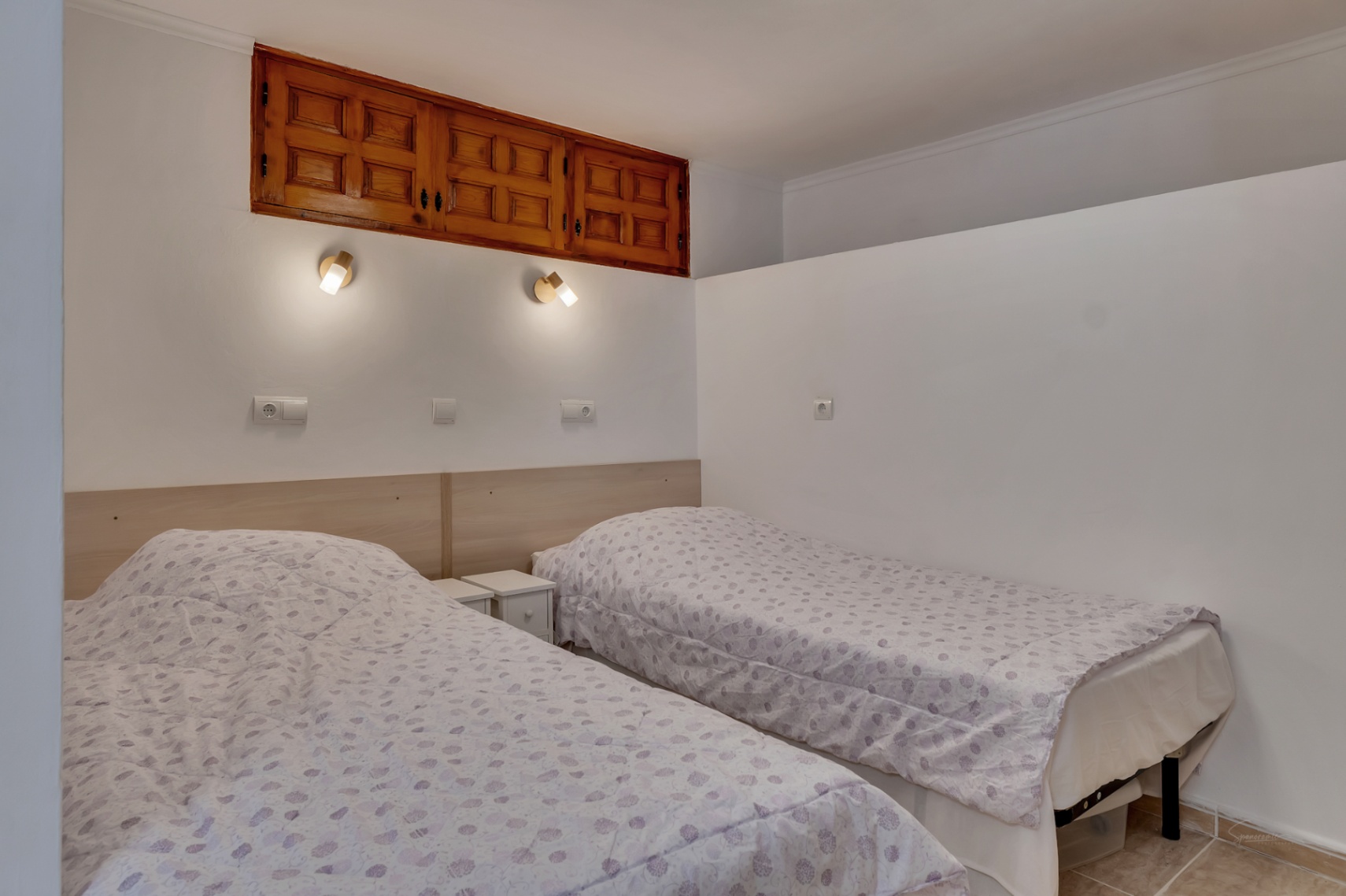 Chambre avec deux lits simples, tête de lit en bois clair et placards muraux traditionnels en bois intégrés au-dessus des liseuses individuelles.