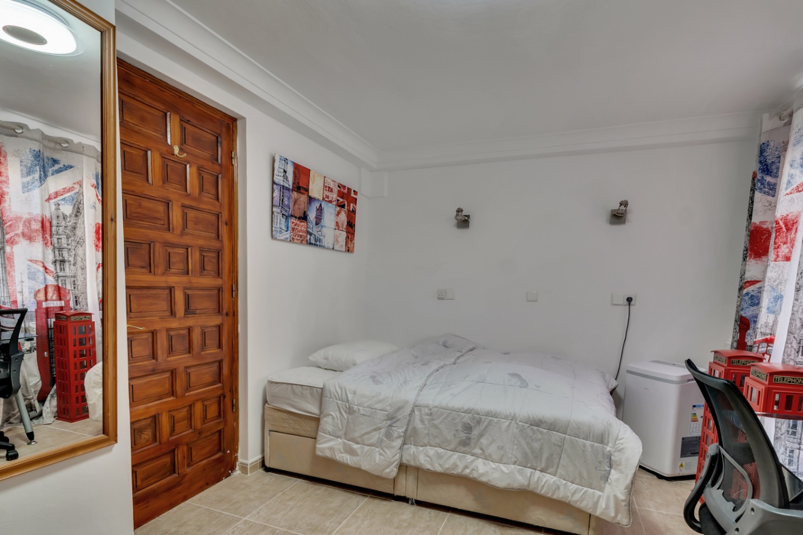 Chambre avec lit double, carrelage au sol et porte en bois massif sculpté. La pièce est équipée d'appliques murales et d'une décoration thématique urbaine.