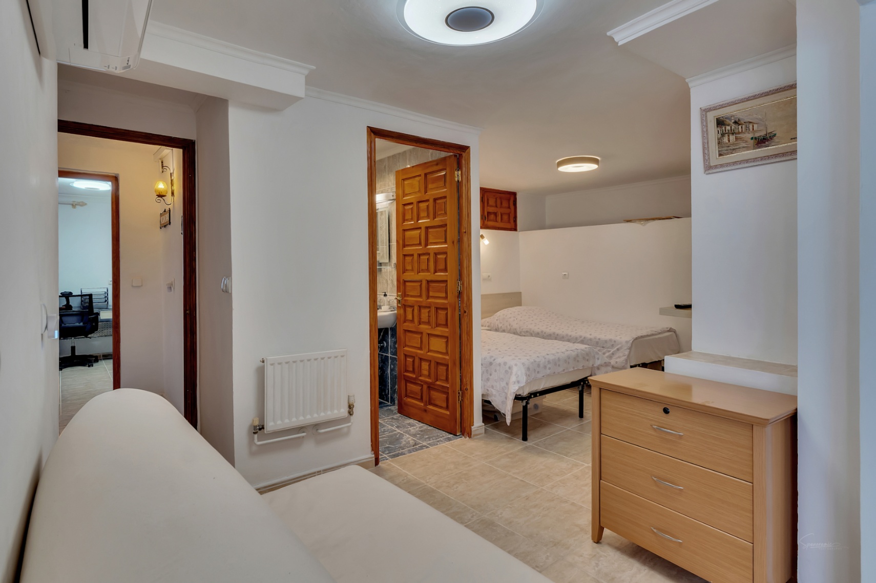 Chambre avec lits jumeaux et salle de bain attenante avec porte en bois sculpté. Comprend un radiateur mural, un sol en céramique et un accès vers un bureau.