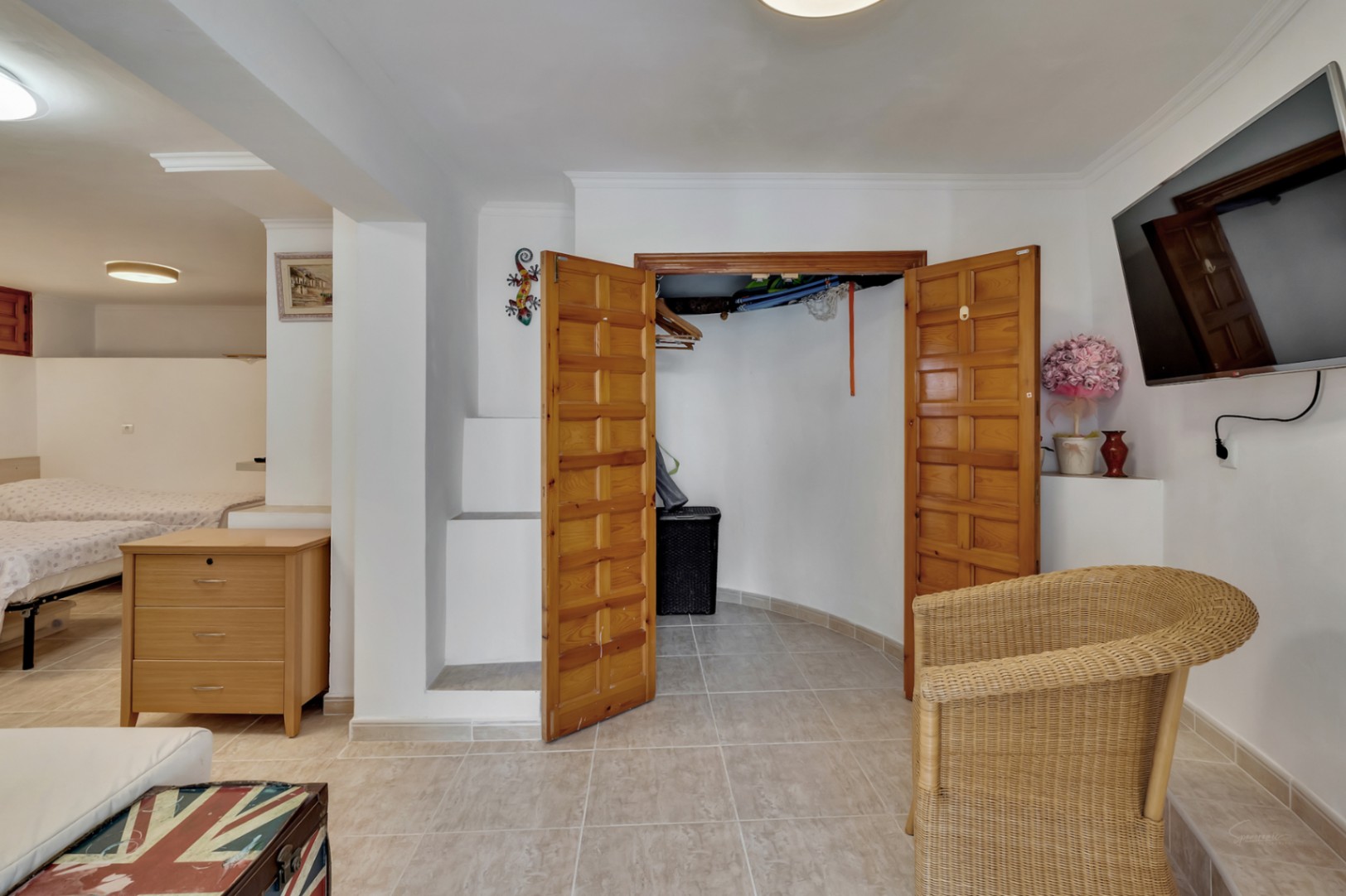 Chambre avec placard intégré en bois, sol en grès cérame et coin salon équipé d'un fauteuil en osier et d'un téléviseur mural.