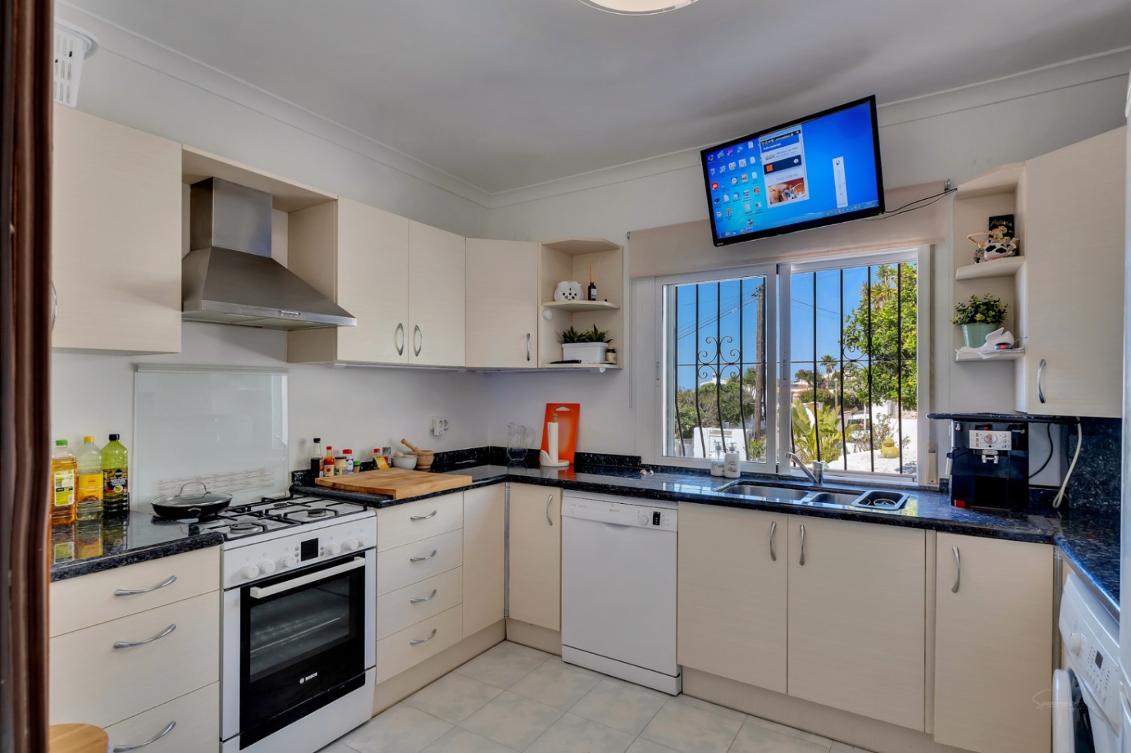 Cuisine fonctionnelle en L avec plans de travail en granit sombre, meubles crème, électroménager blanc et fenêtre avec vue sur le jardin et télévision murale.