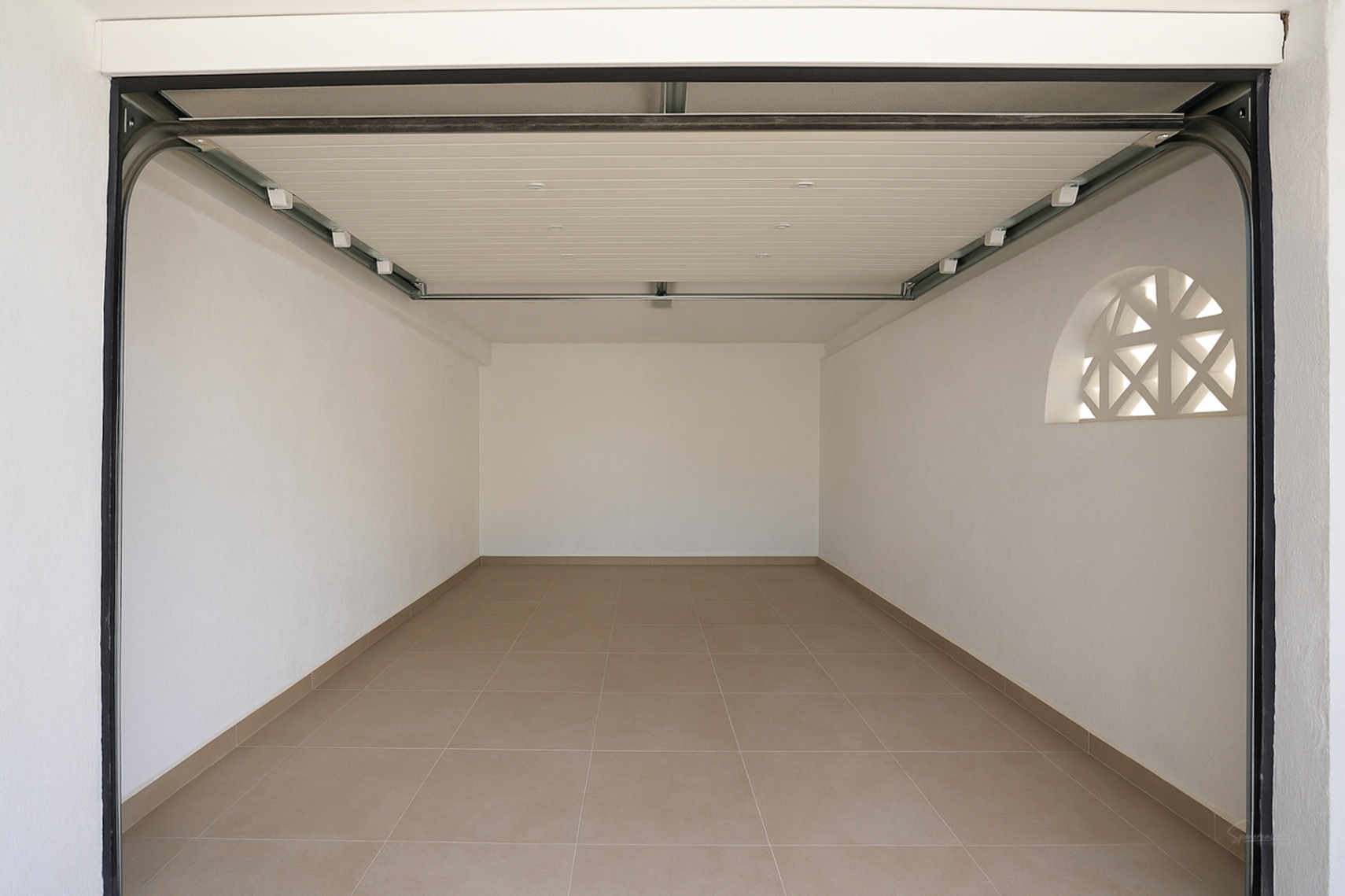 Garage privé avec sol en carrelage, murs blancs et porte sectionnelle automatique. Une fenêtre décorative en arche apporte de la lumière naturelle.