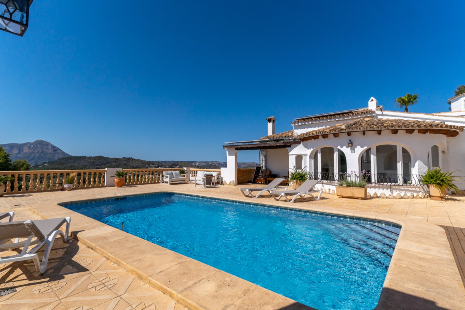 Piscine extérieure entourée d'une vaste terrasse en pierre avec vue panoramique sur le Montgó. La villa blanche présente des arches traditionnelles et des panneaux solaires.