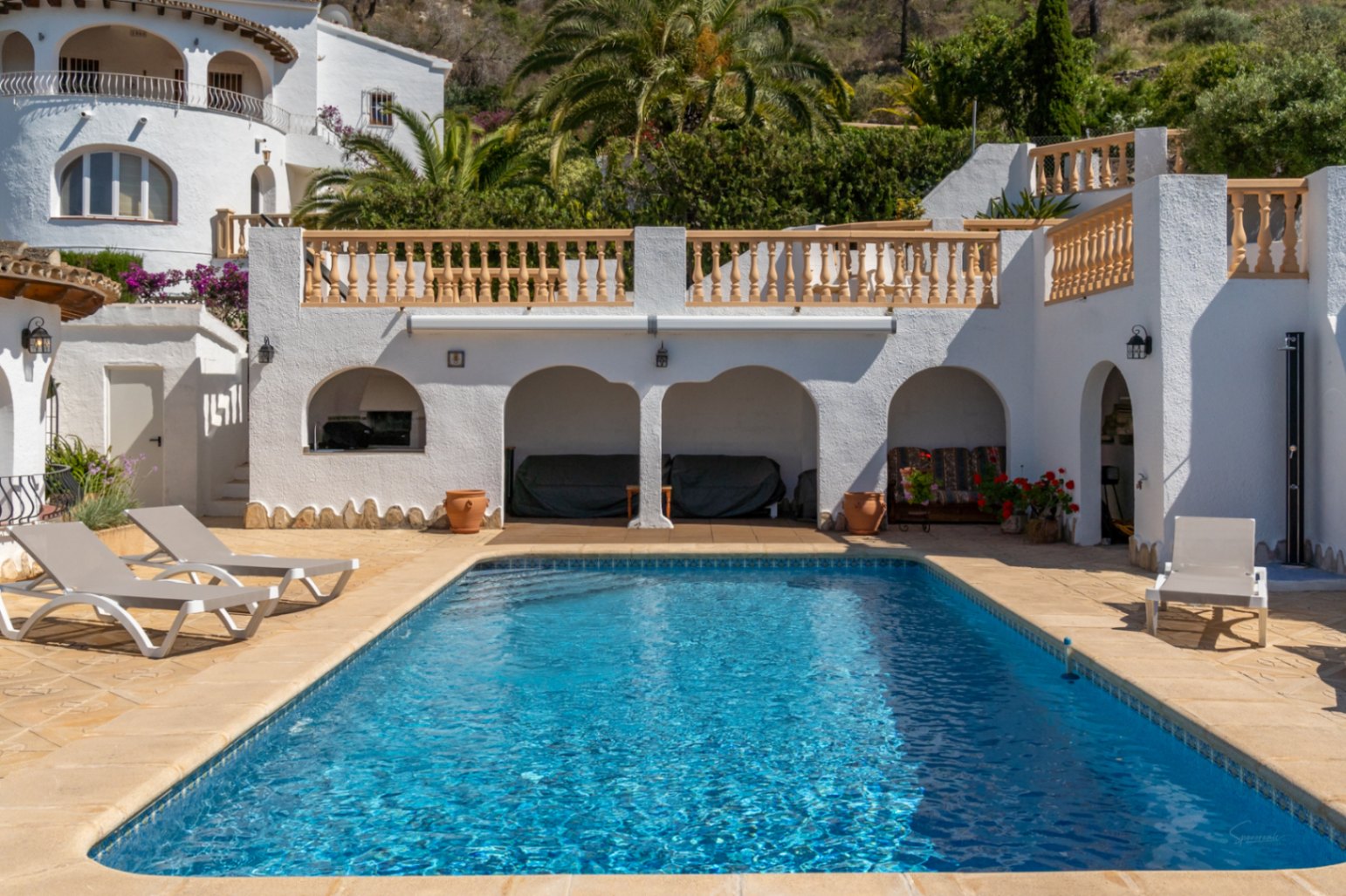 Piscine rectangulaire sur une terrasse en pierre avec transats, caractérisée par des porches voûtés et des balustrades blanches dans un cadre méditerranéen.