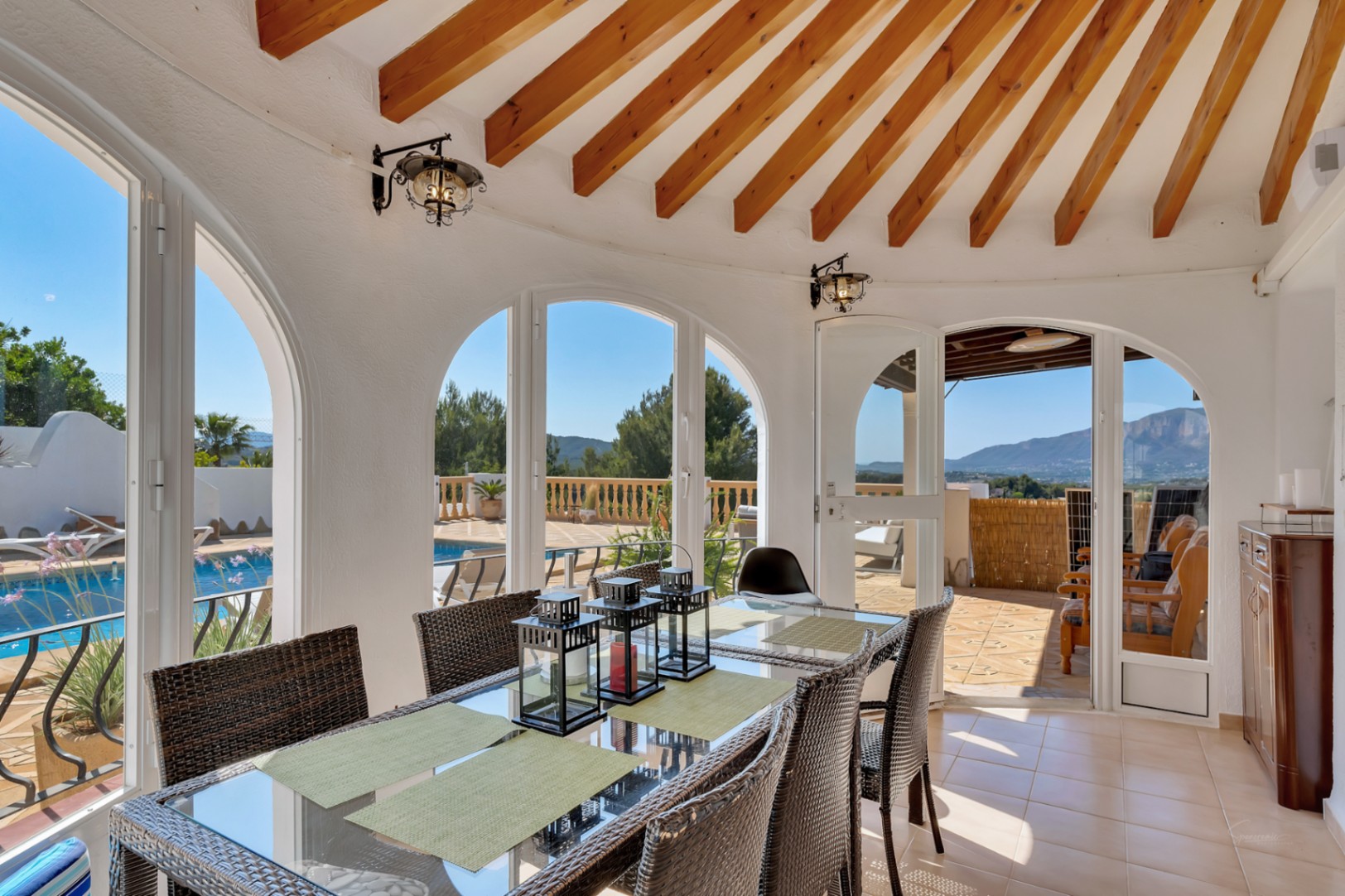 Salle à manger couverte avec poutres apparentes en éventail, grandes fenêtres cintrées offrant une vue sur la piscine et le massif du Montgó.