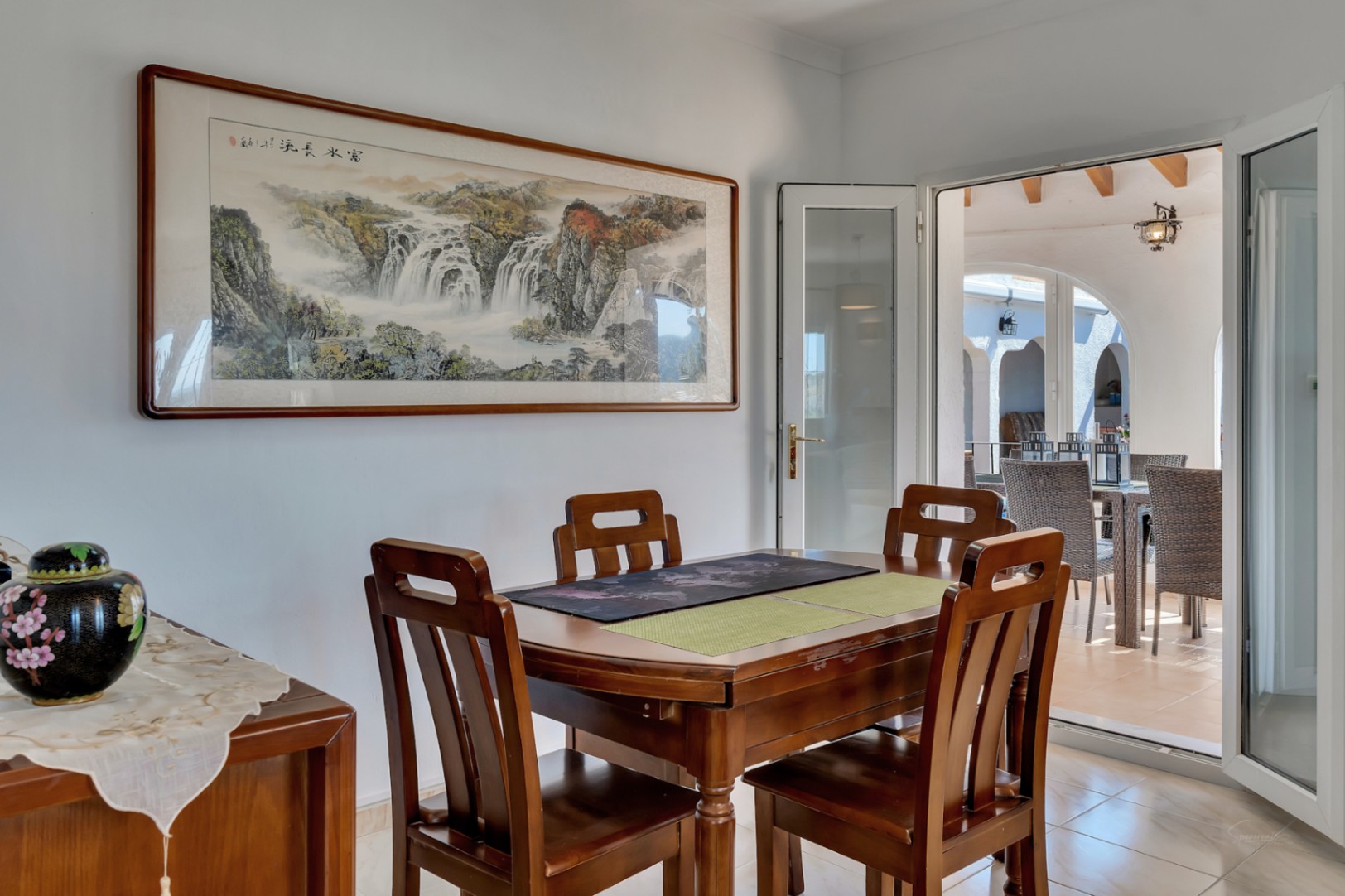 Salle à manger intérieure avec mobilier en bois et accès direct à la terrasse. La pièce est décorée d'un grand tableau paysager et baignée de lumière naturelle.
