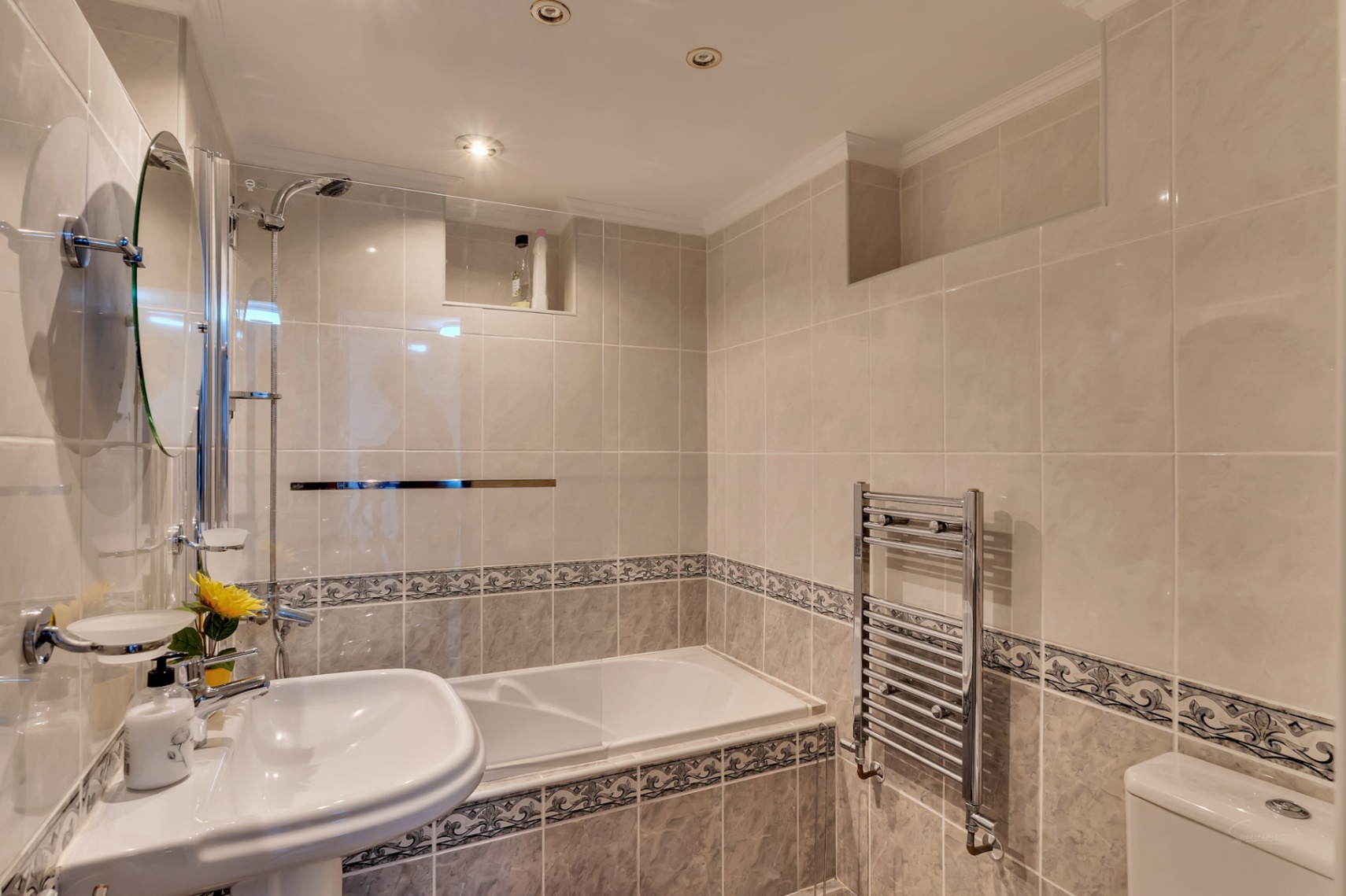 Salle de bain avec baignoire et paroi vitrée, carrelage crème avec frise décorative, sèche-serviettes chromé et éclairage encastré au plafond.