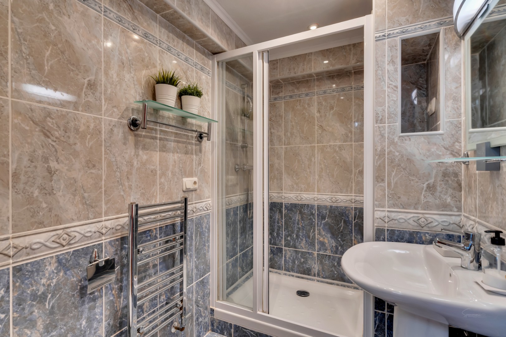 Salle de bain avec douche à paroi blanche, carrelage effet marbre beige et bleu, radiateur sèche-serviettes chromé et lavabo sur colonne.