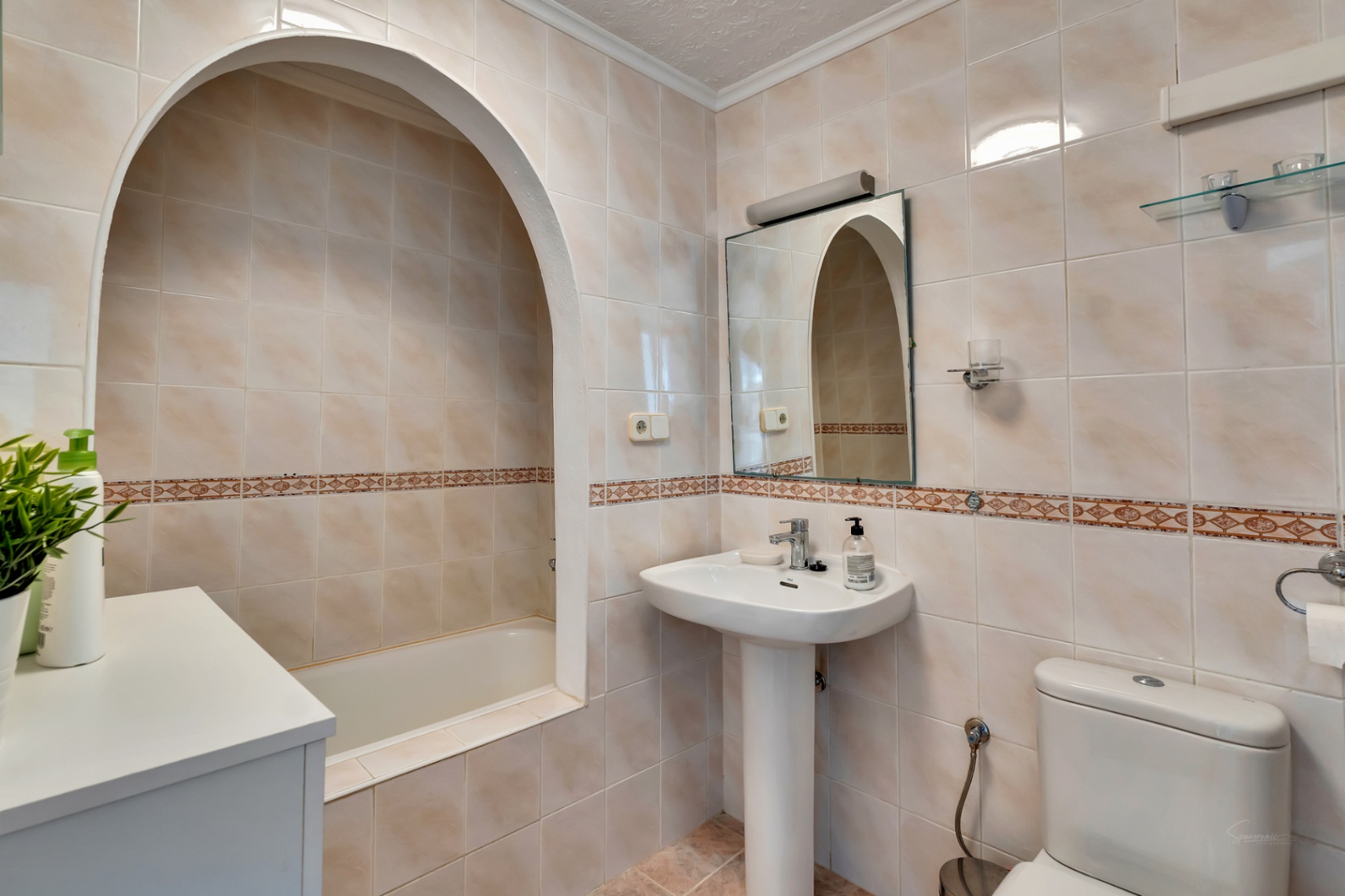 Salle de bain classique avec baignoire encastrée dans une alcôve voûtée, lavabo sur colonne et carrelage mural beige avec frise décorative.