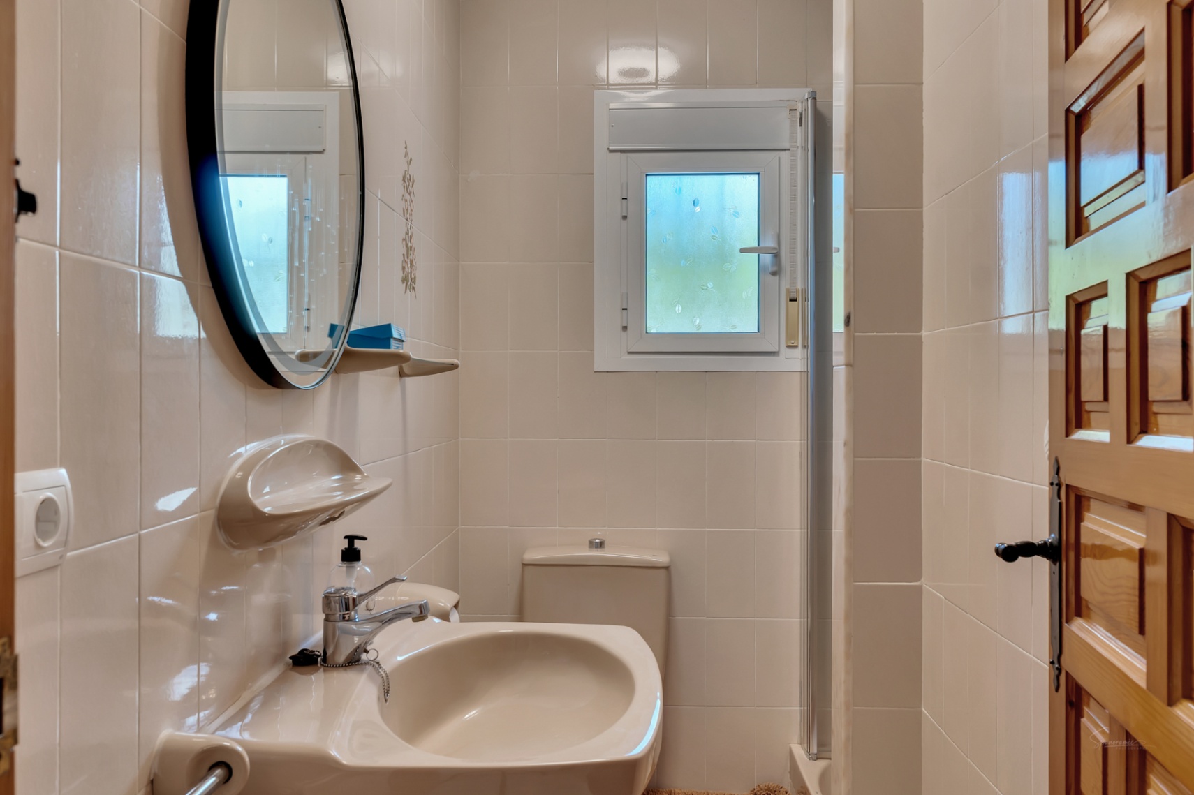 Salle de bain fonctionnelle avec carrelage crème, lavabo assorti, miroir circulaire à cadre noir et petite fenêtre oscillo-battante pour la ventilation.