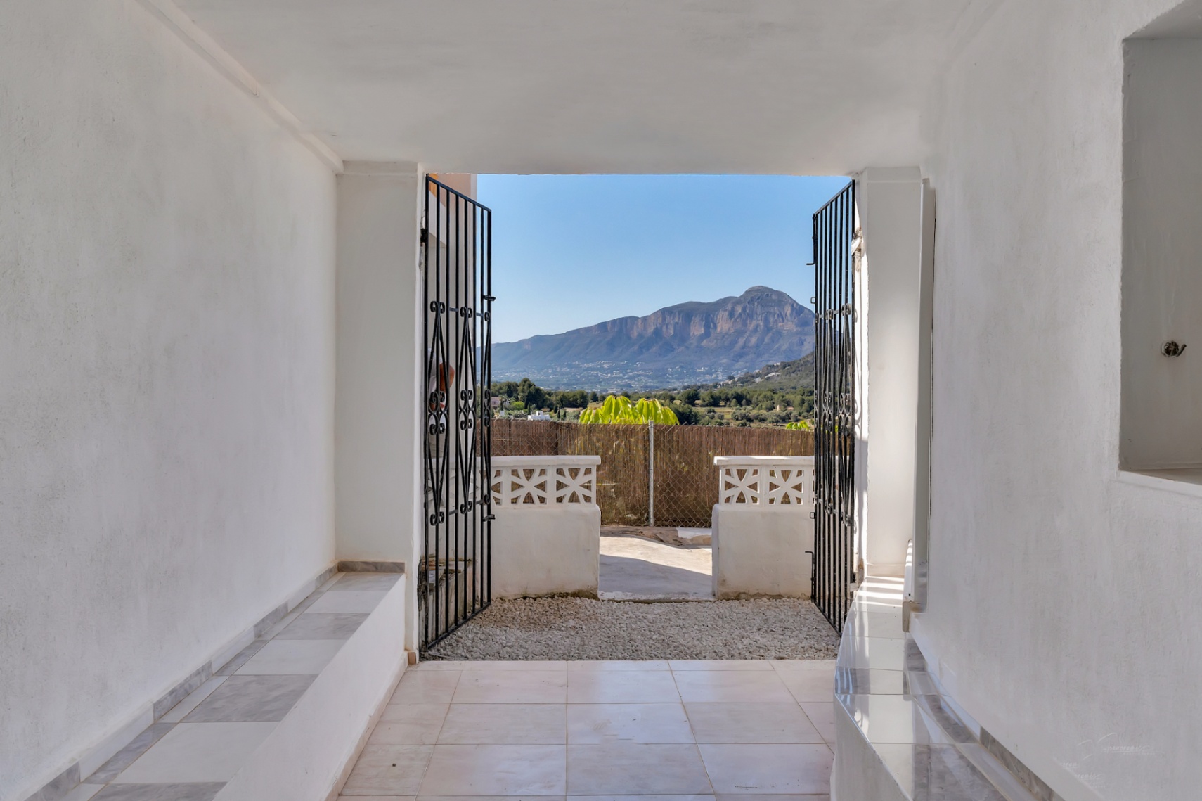 Vue panoramique sur le Montgó depuis un porche d'entrée aux murs blancs, avec sol en carrelage et grilles en fer forgé ouvrant sur le paysage méditerranéen.