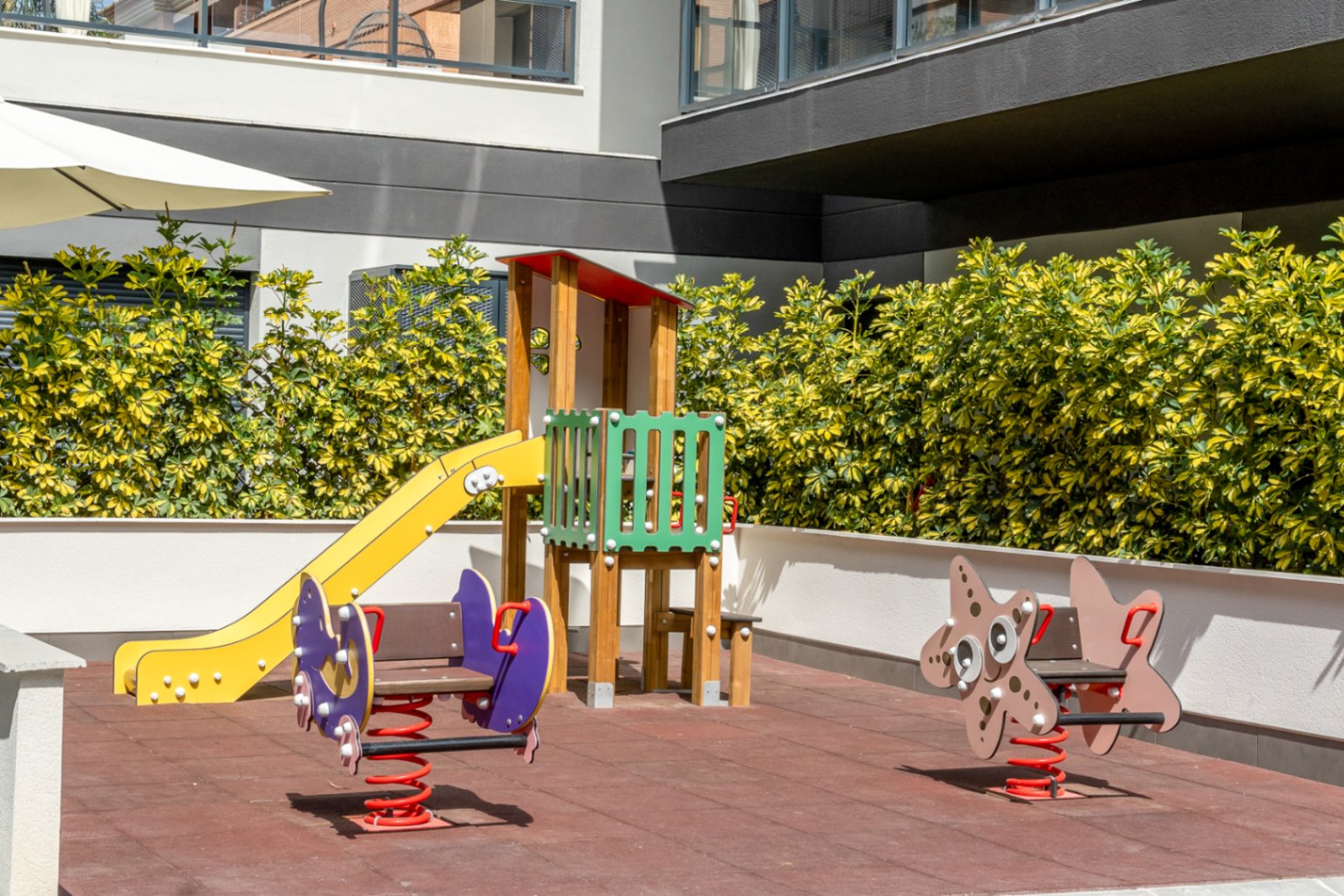 Aire de jeux pour enfants avec sol de sécurité, toboggan en bois et jeux à ressort, entourée de haies soignées au sein du complexe résidentiel.