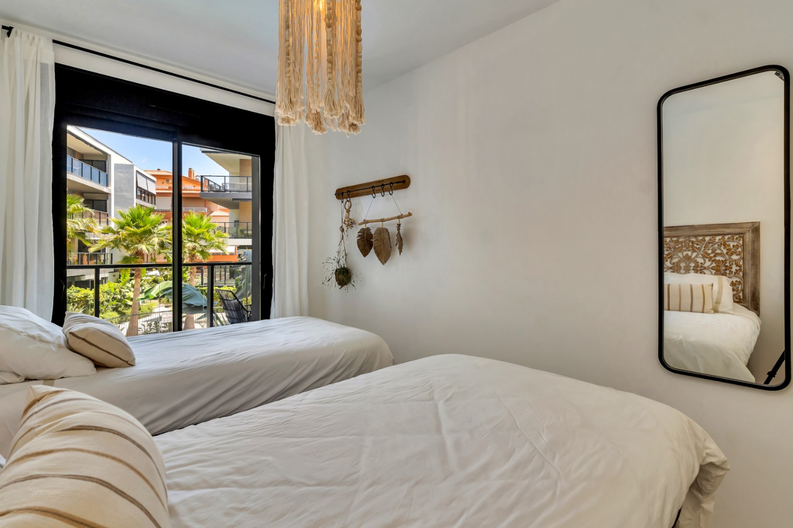 Chambre avec deux lits simples, luminaire en macramé et miroir vertical. La grande baie vitrée donne sur les jardins et palmiers de la copropriété.