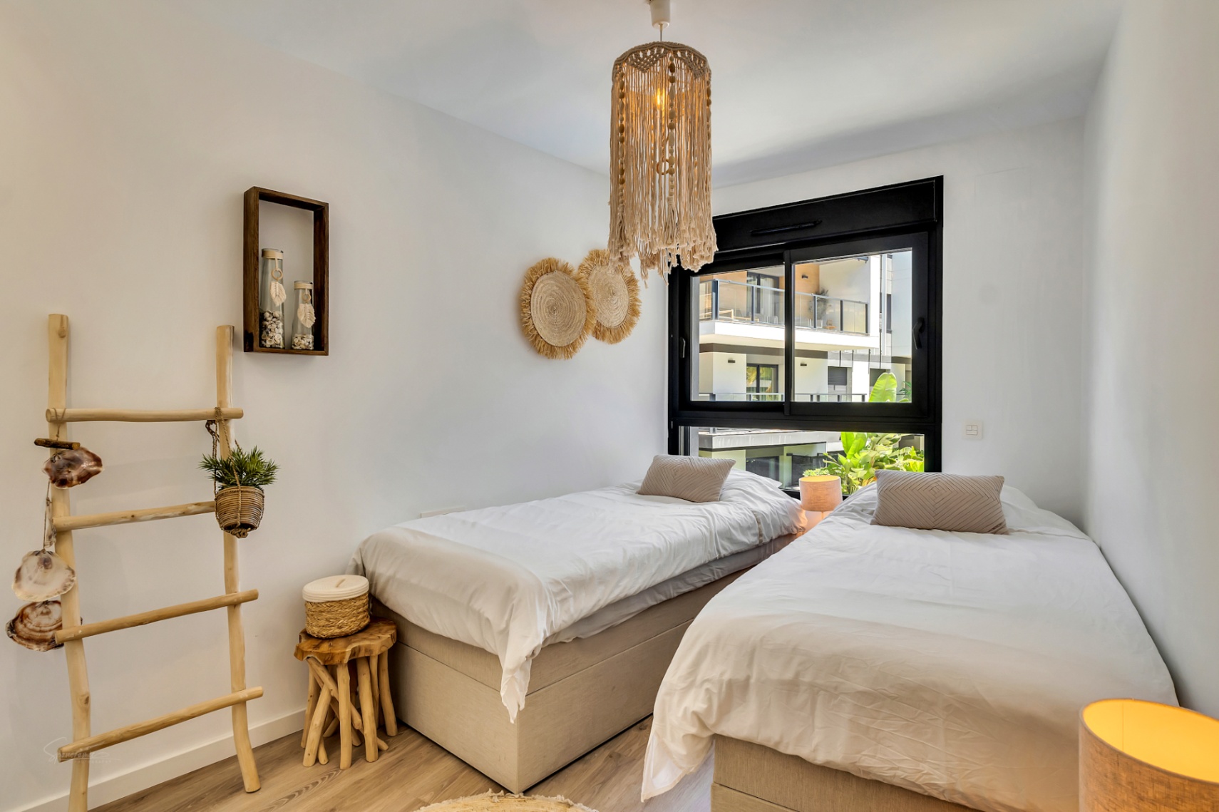 Chambre lumineuse avec deux lits simples, décorée d'éléments en bois naturel, d'une suspension en macramé et d'une fenêtre à cadre noir offrant une vue extérieure.