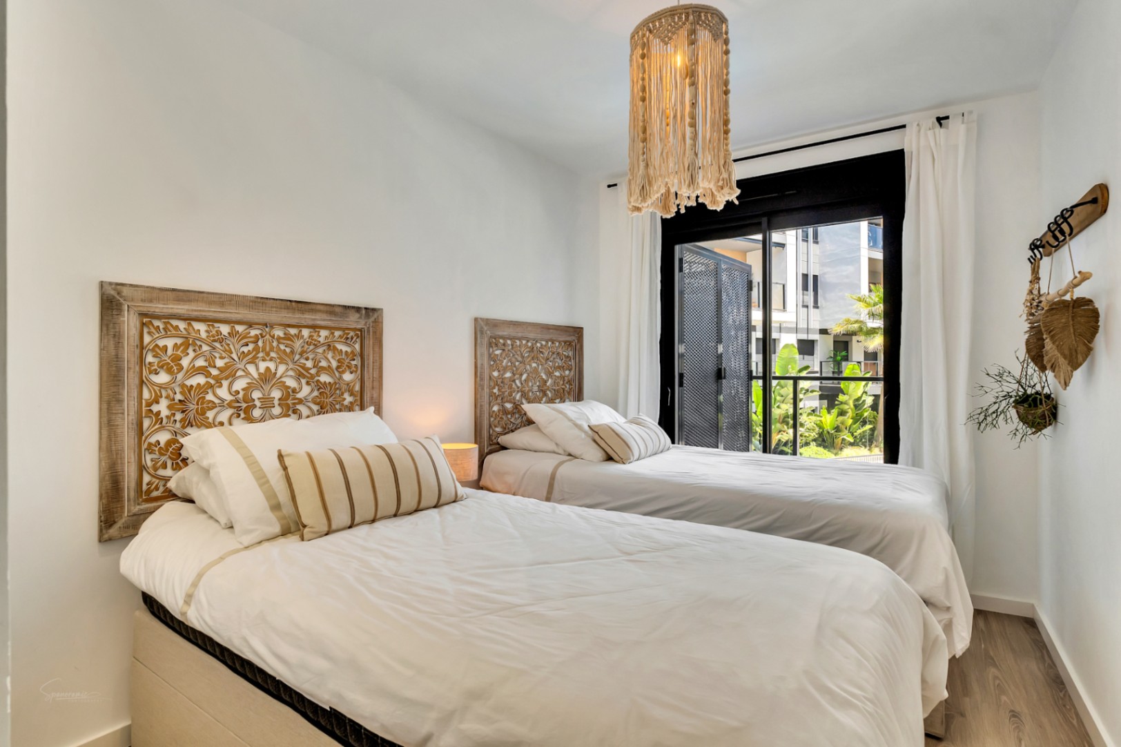 Chambre lumineuse avec deux lits simples, têtes de lit en bois sculpté et suspension en macramé. Une grande baie vitrée donne sur une terrasse avec vue sur les jardins.