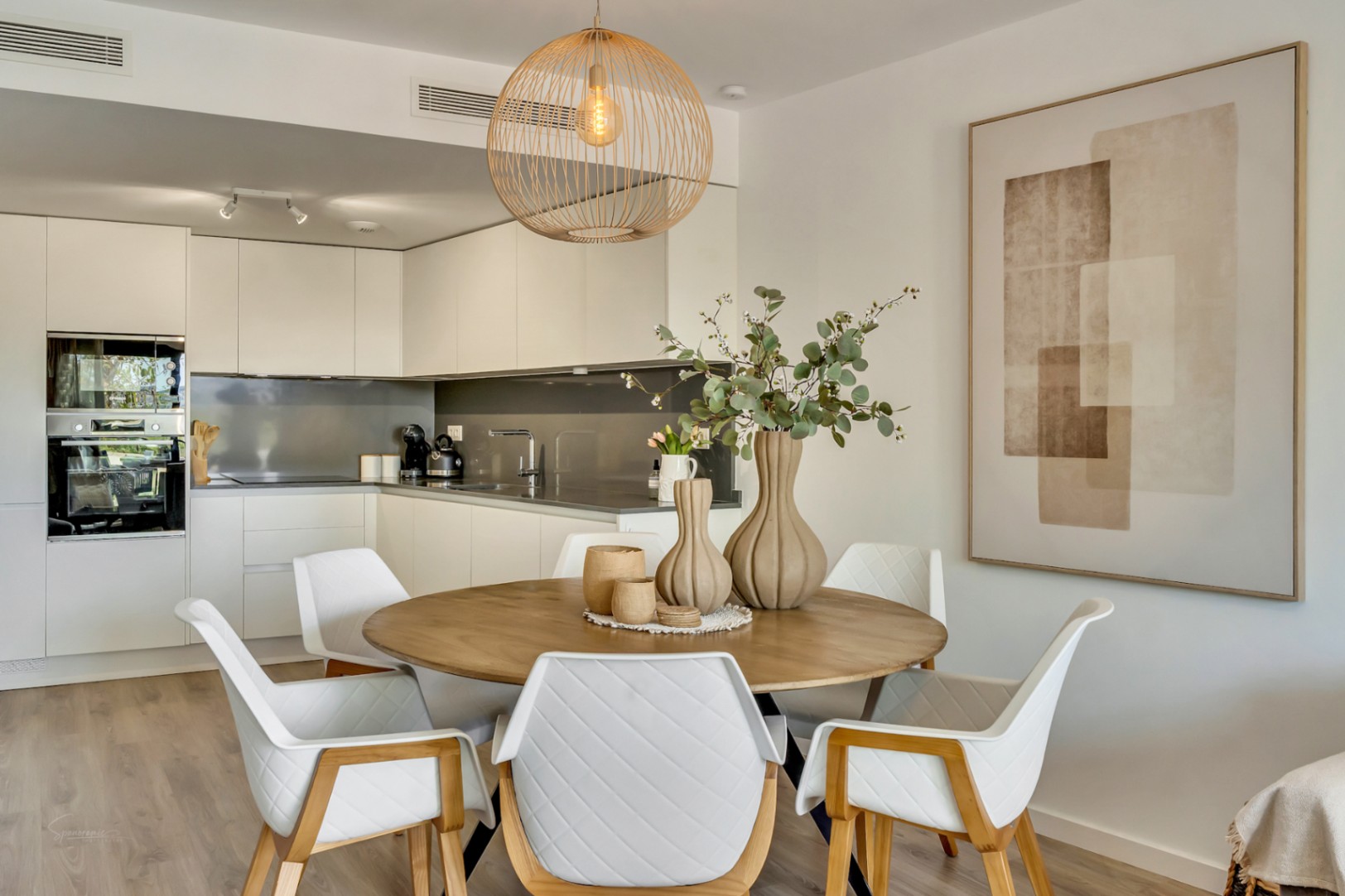 Coin repas à aire ouverte avec table ronde en bois, chaises matelassées et suspension en osier, s'ouvrant sur une cuisine moderne aux finitions épurées.