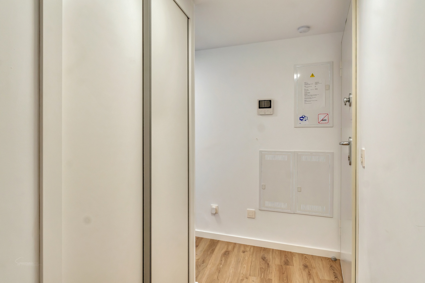Couloir d'entrée avec parquet clair, grand placard intégré blanc et panneaux de contrôle techniques encastrés pour un design épuré et fonctionnel.