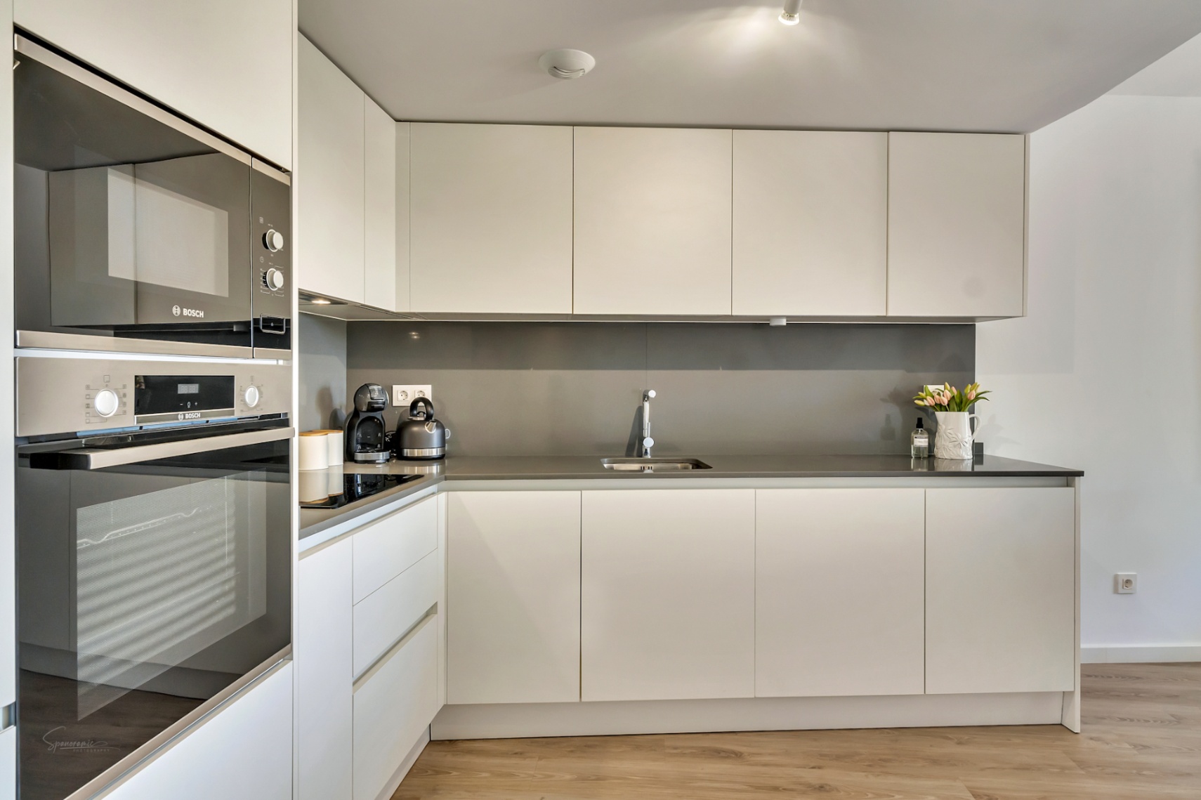 Cuisine minimaliste moderne avec meubles blancs sans poignées, plan de travail gris et électroménagers Bosch intégrés sur un sol en bois clair.
