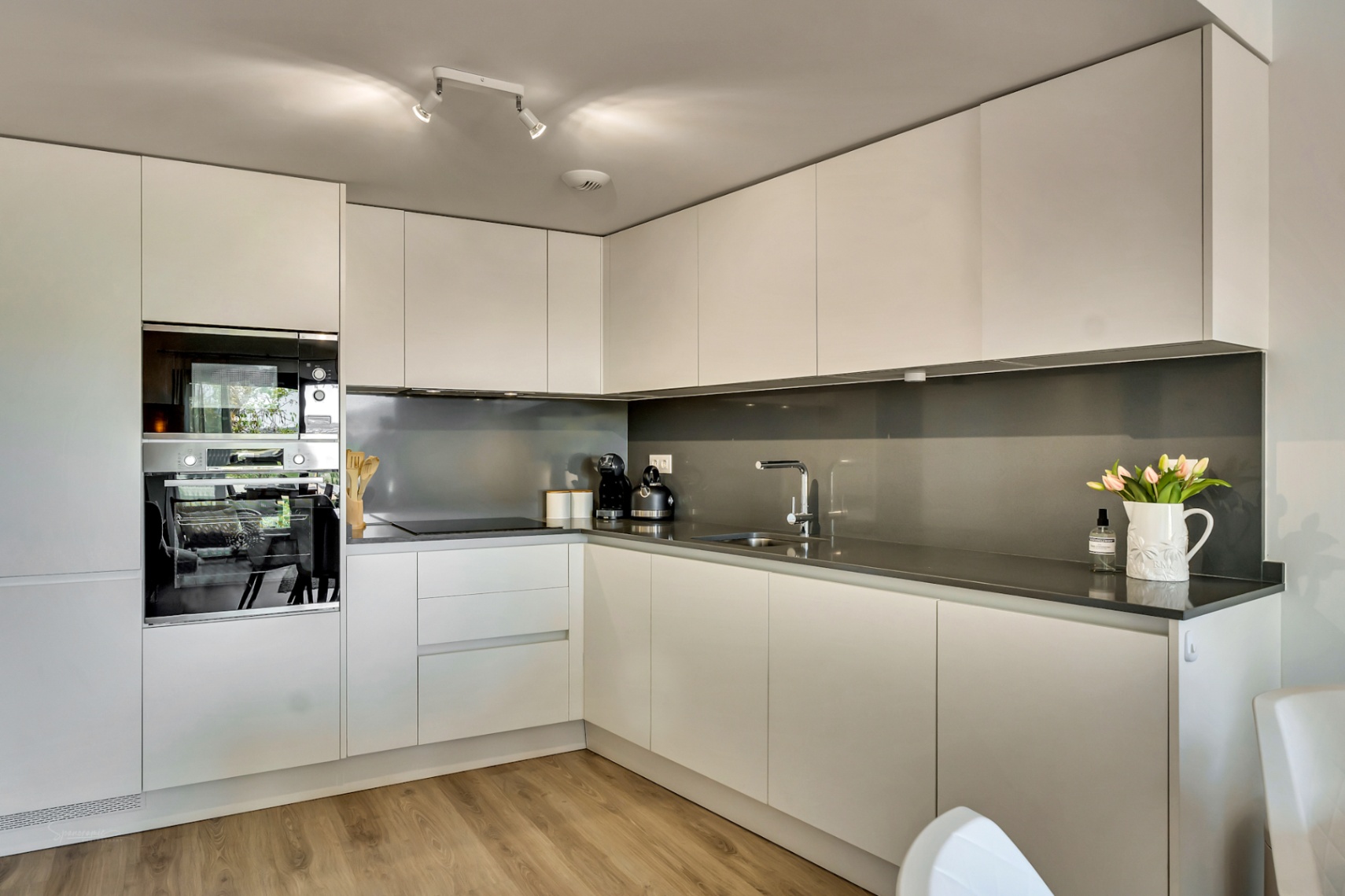 Cuisine moderne en L avec des meubles blancs sans poignées, un plan de travail en quartz gris foncé et des appareils électroménagers intégrés de haute qualité.