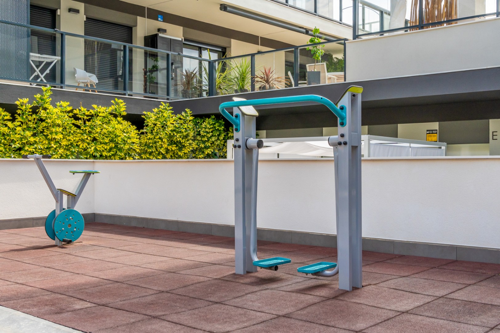 Espace de sport en plein air équipé de machines de fitness sur revêtement de sécurité, entouré de haies verdoyantes et de la façade moderne de la résidence.