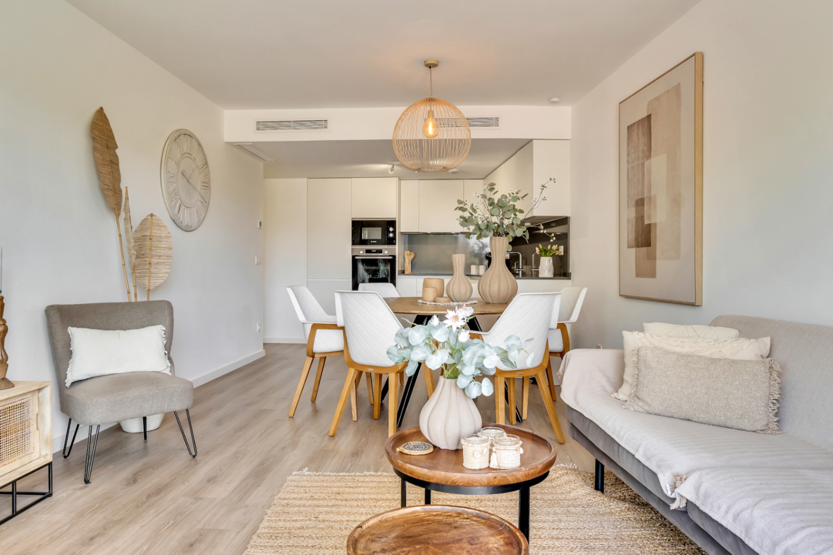 Espace de vie ouvert comprenant un salon et une salle à manger aux tons neutres, avec parquet clair et une cuisine moderne équipée de meubles blancs minimalistes.
