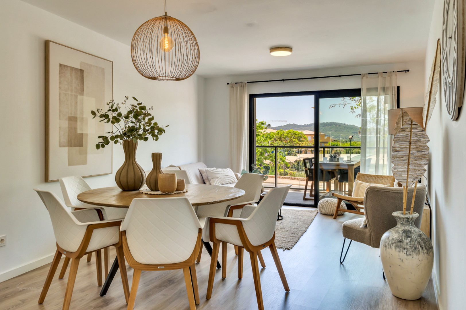Espace de vie ouvert comprenant une salle à manger avec table ronde en bois et un salon lumineux s'ouvrant sur une terrasse avec vue sur les collines.