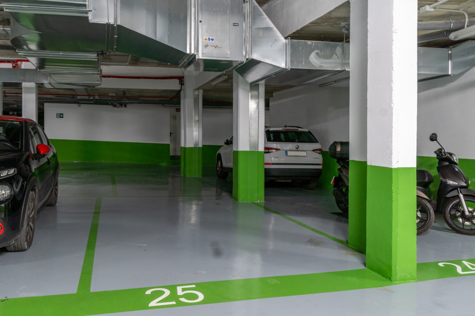 Place de parking souterraine numérotée avec sol en résine grise et marquages verts. Comprend des piliers en béton et des conduits de ventilation apparents.