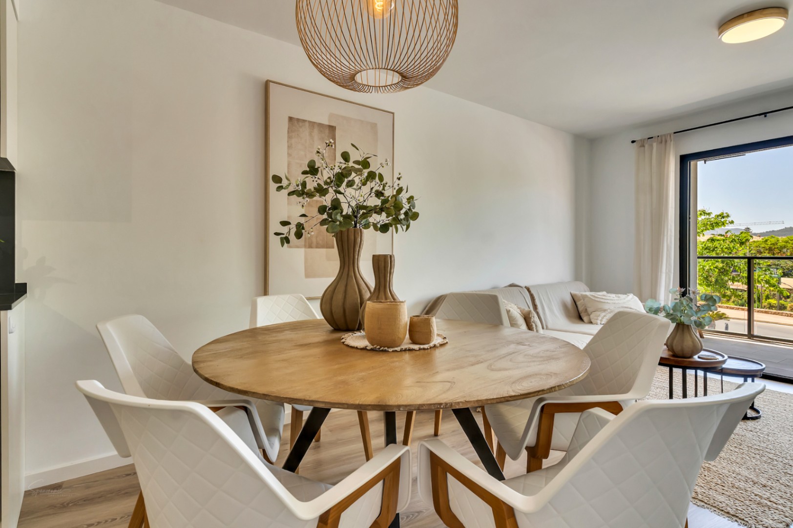 Salle à manger ouverte avec table ronde en bois, chaises matelassées et suspension en rotin, s'ouvrant sur un salon lumineux et un balcon avec vue dégagée.