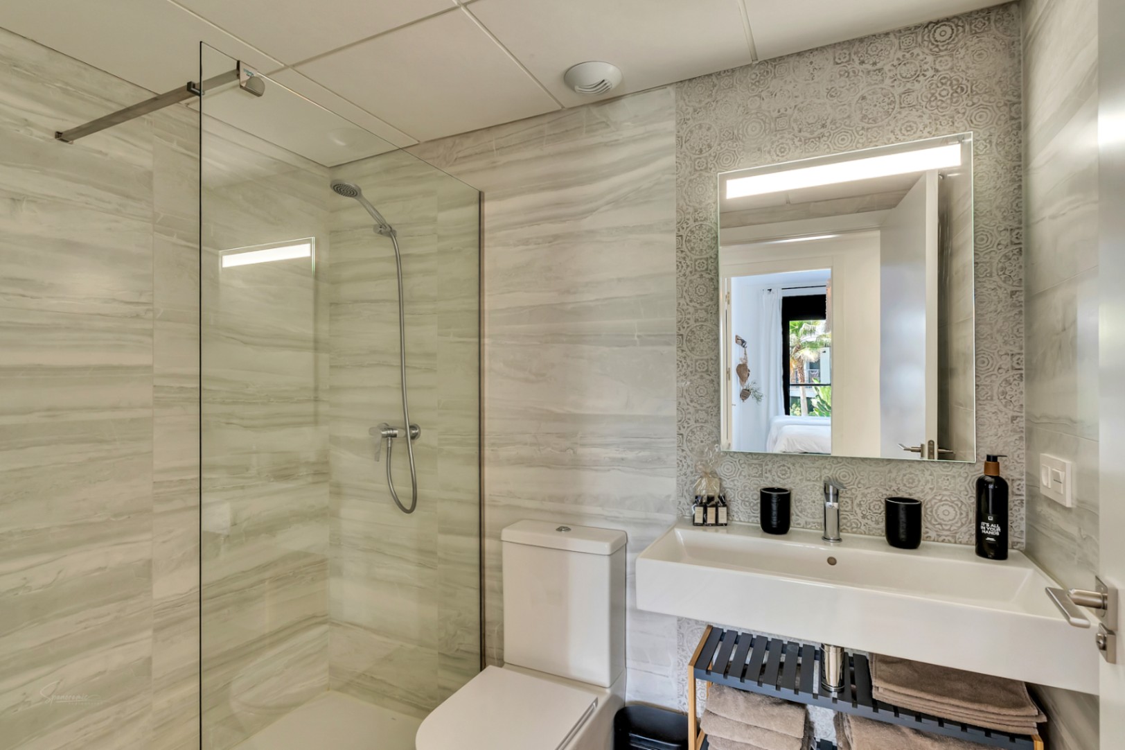 Salle de bain moderne avec douche à l'italienne, paroi en verre et carrelage effet marbre. Elle dispose d'un lavabo suspendu sur un mur décoratif et d'un miroir rétroéclairé.