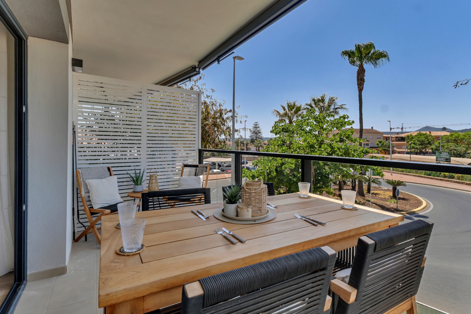 Terrasse couverte avec table à manger en bois, chaises en corde noire et vue dégagée sur les palmiers et les environs de Jávea.