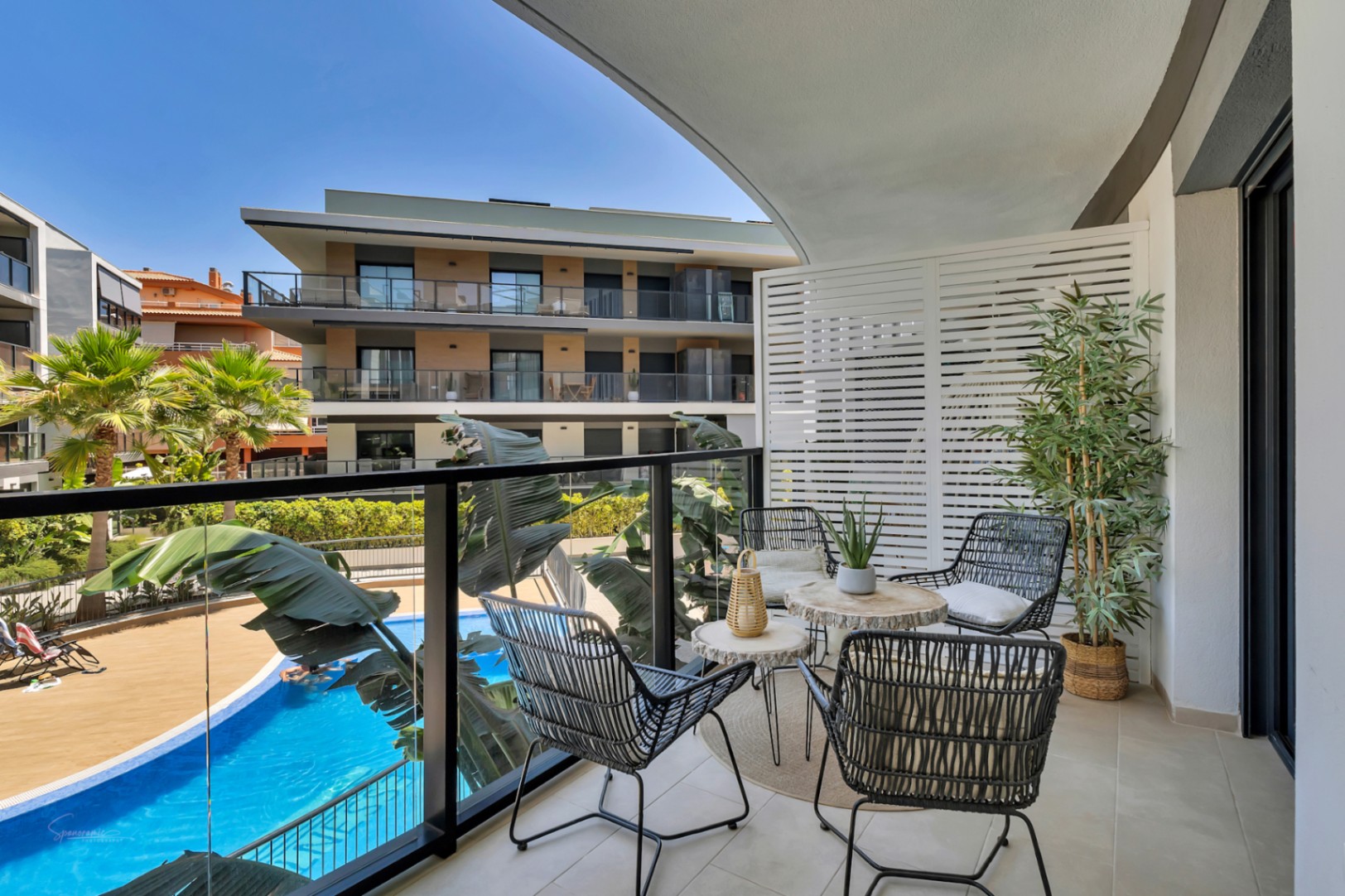 Terrasse privée avec mobilier moderne, garde-corps en verre et vue dégagée sur la piscine commune et les jardins paysagers de la résidence.