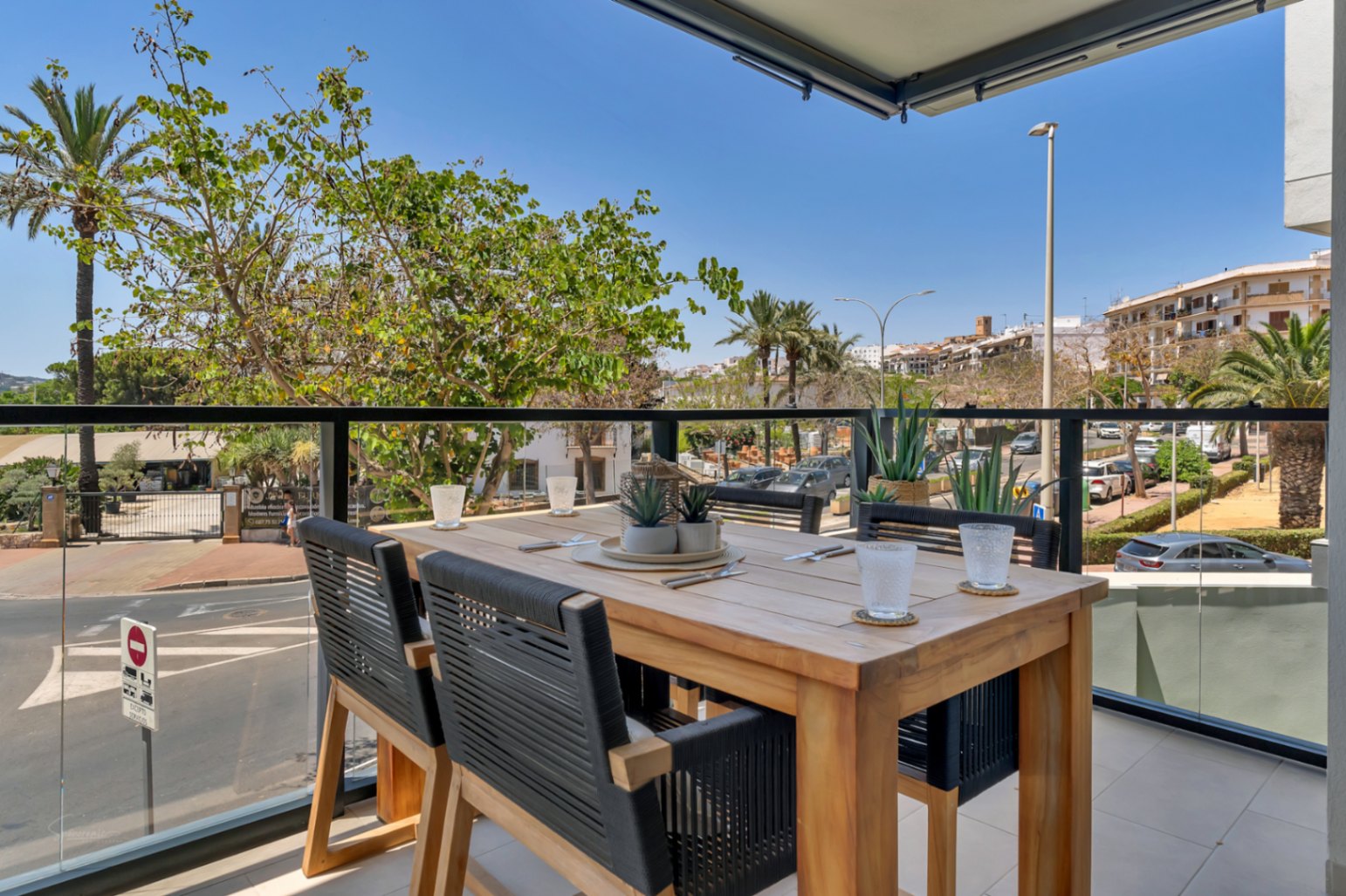 Terrasse privée avec une table à manger en bois, des chaises en corde tressée et un garde-corps en verre offrant une vue dégagée sur les environs urbains.