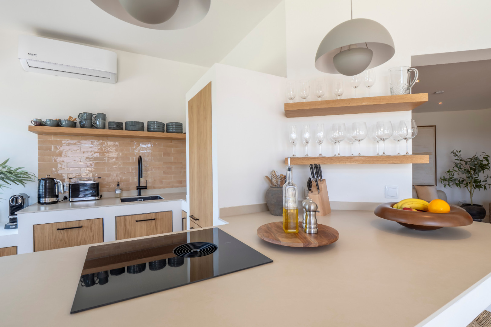 Détail de la cuisine avec comptoir beige, plaque vitrocéramique noire et étagères en bois clair avec verrerie.