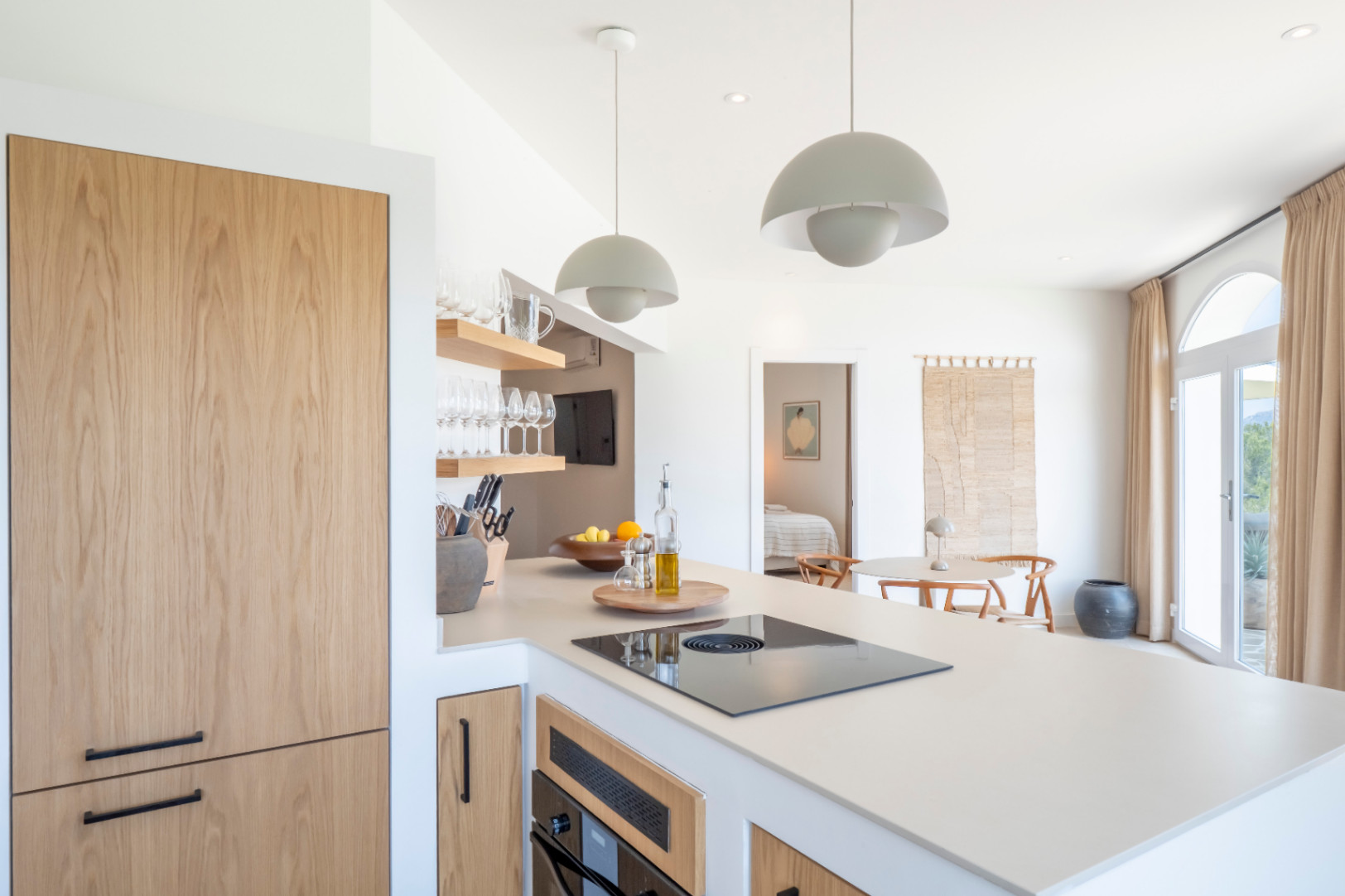 Détail de la cuisine avec îlot, électroménagers à panneaux en bois clair et étagères ouvertes avec verrerie.
