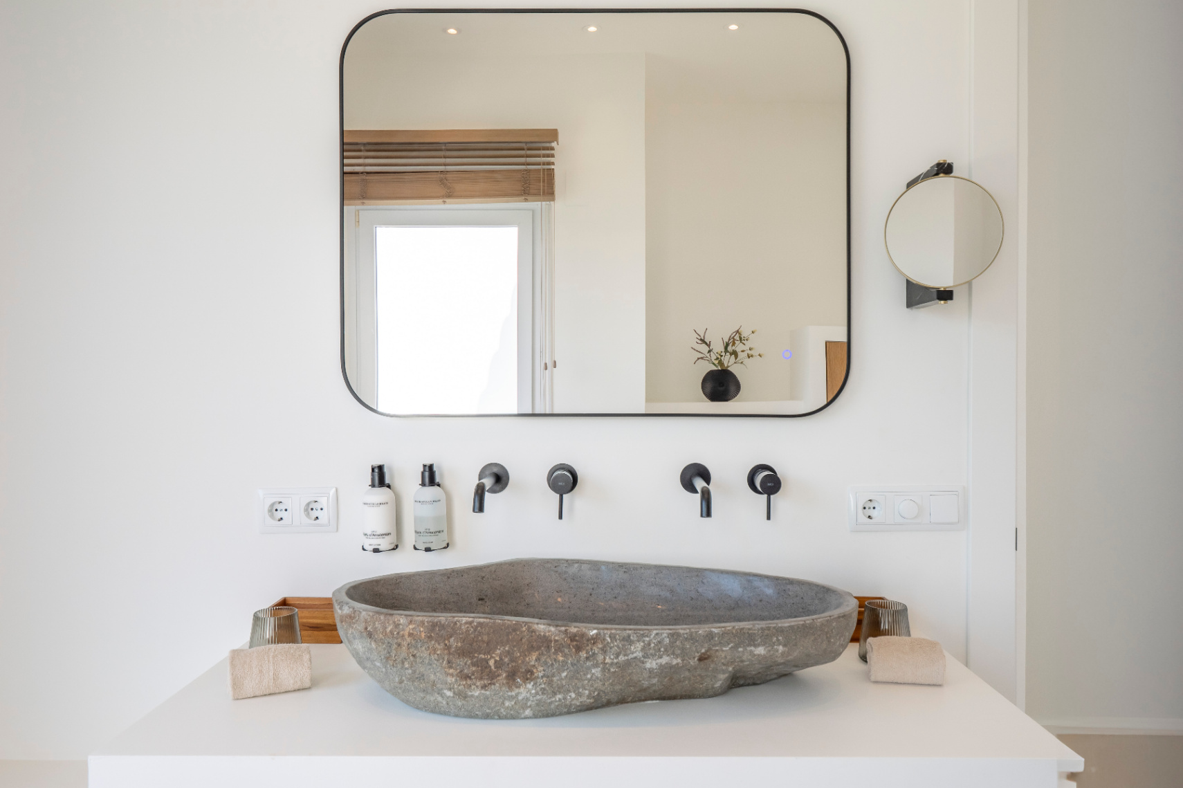 Détail de salle de bain avec lavabo en pierre naturelle et robinetterie noire mate. Miroir rectangulaire avec éclairage intégré.