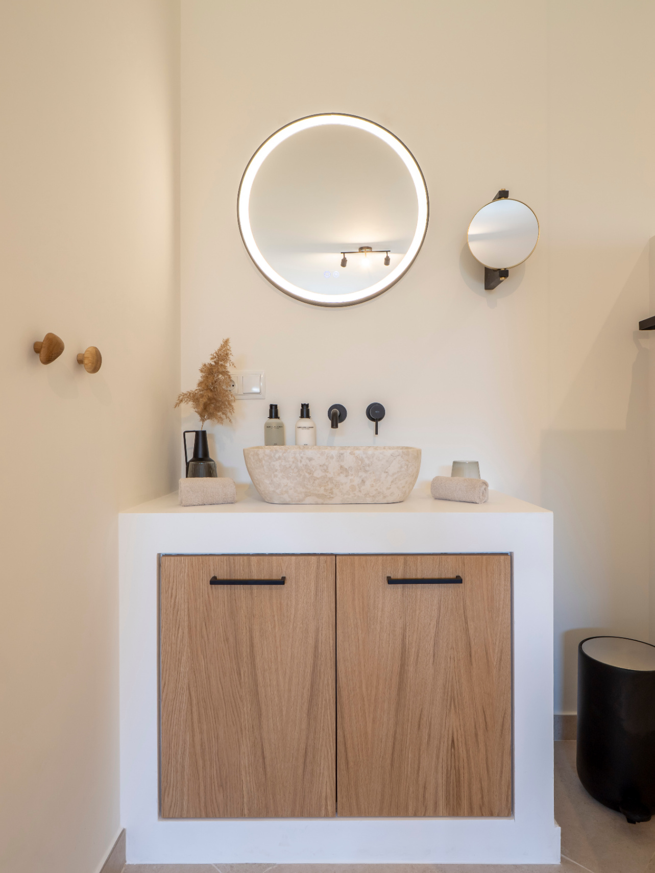 Détail de salle de bain avec vasque en pierre naturelle, miroir circulaire rétroéclairé et meuble en bois clair à poignées noires.