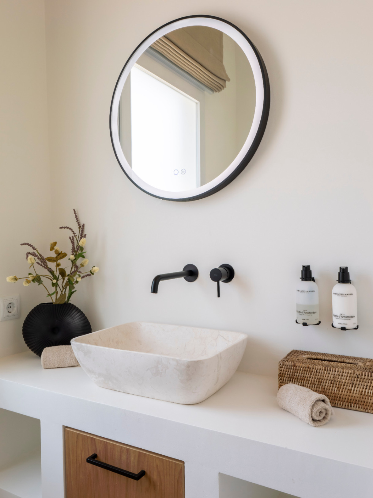 Détail de salle de bain avec vasque en pierre, robinetterie noire mate et miroir rond à éclairage intégré.