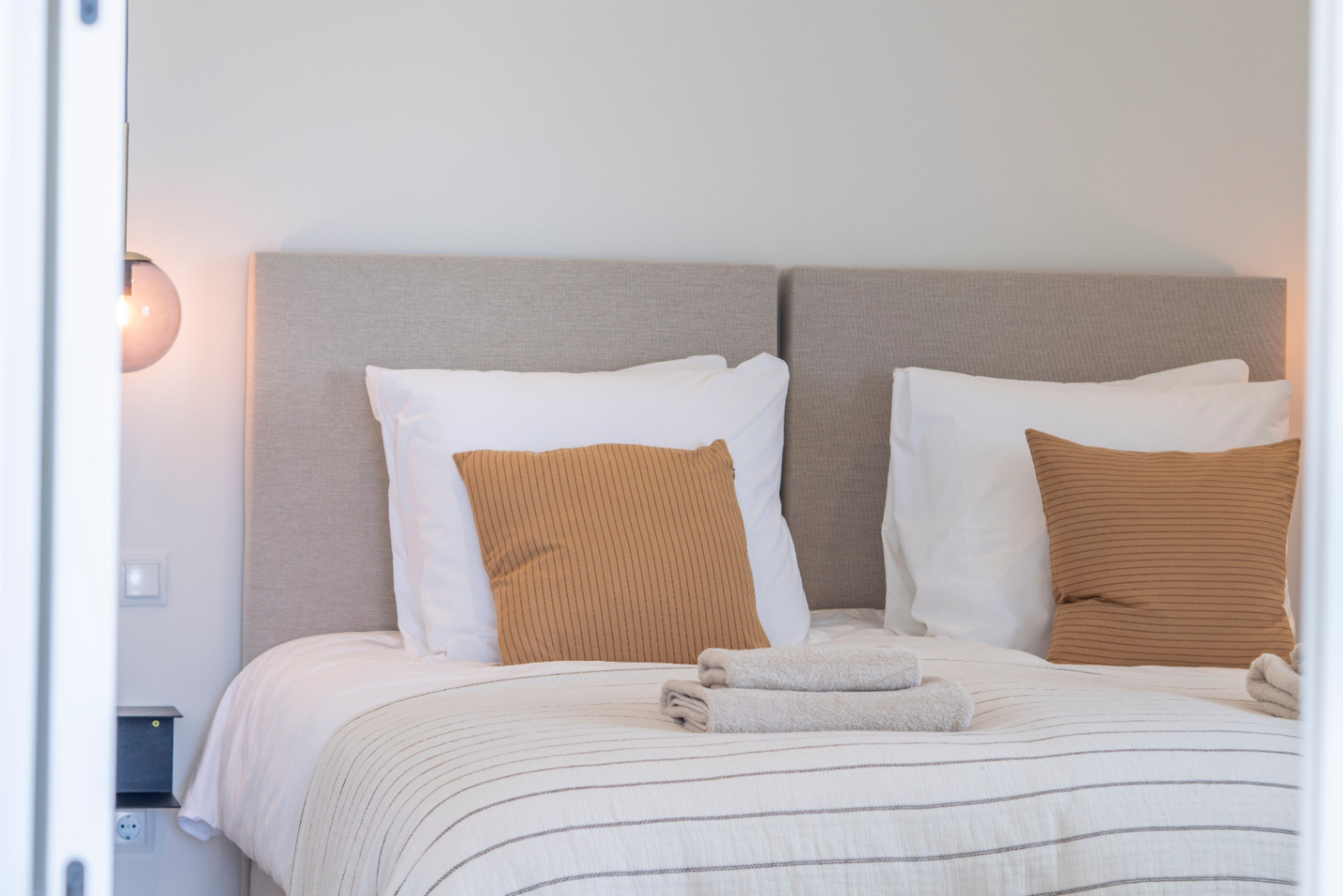 Détail d'une chambre avec tête de lit rembourrée, coussins moutarde et linge de lit rayé.