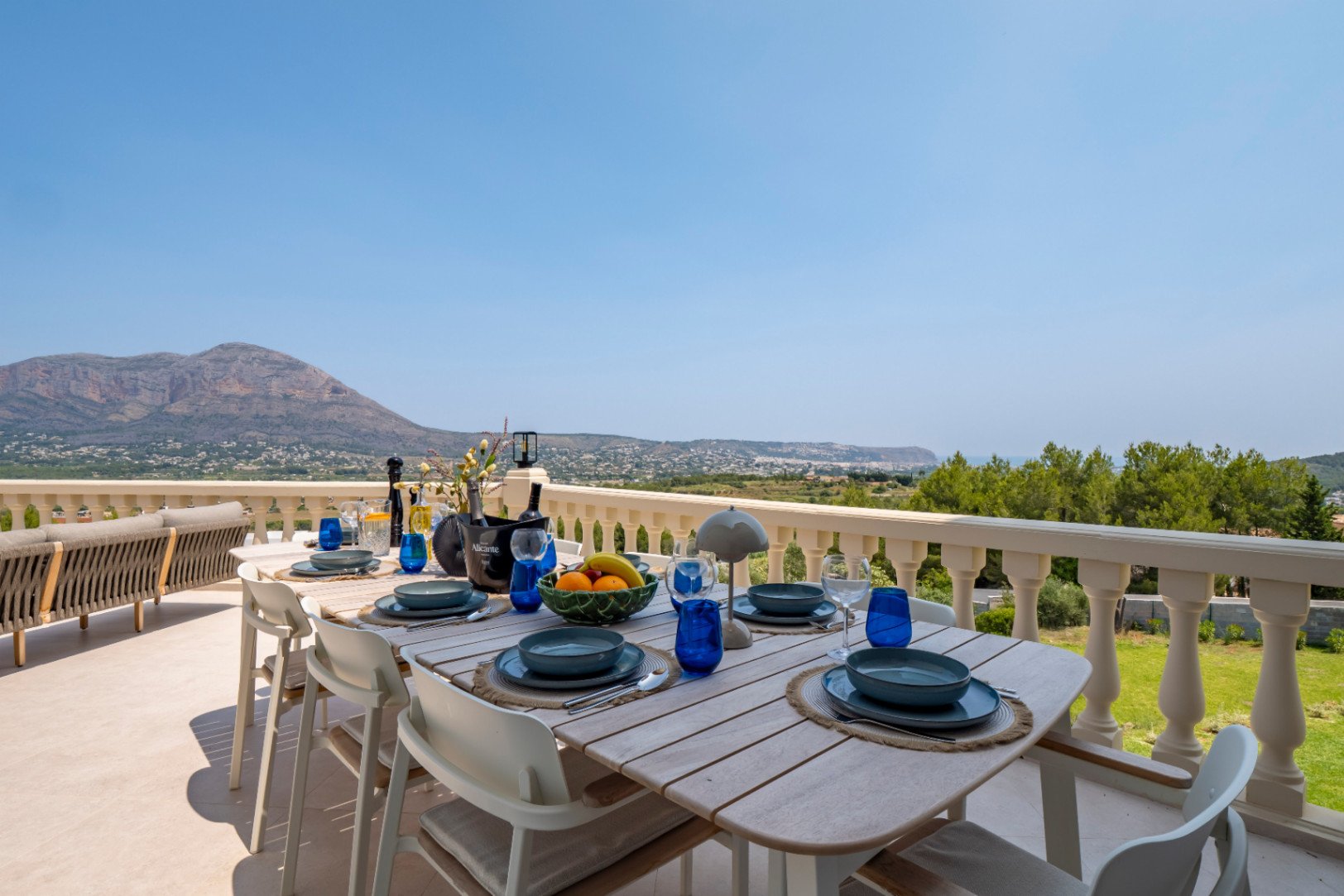 Salle à manger extérieure avec table en bois et vue panoramique sur la montagne et la côte depuis la terrasse.