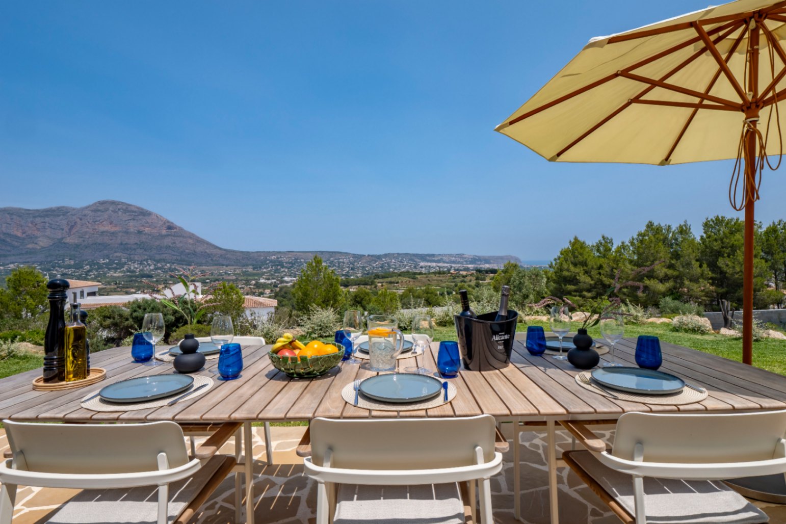 Salle à manger extérieure avec table en bois, vaisselle bleue et vue panoramique sur la montagne à Jávea.