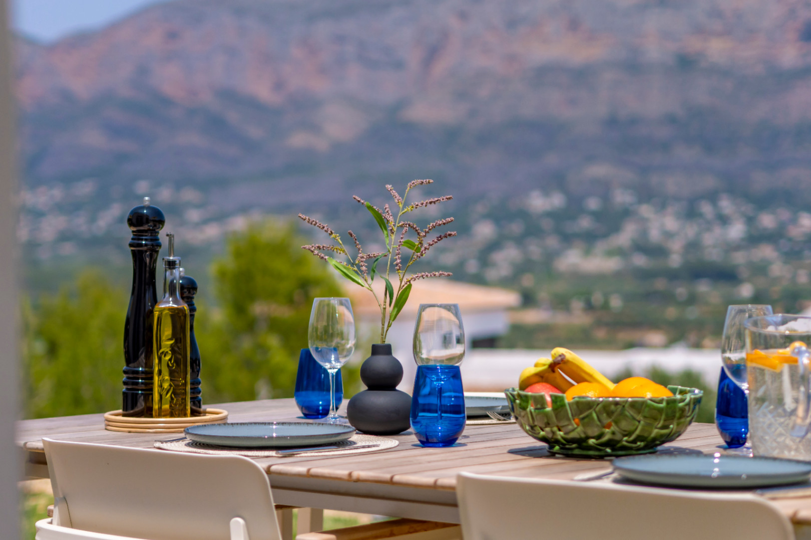 Salle à manger extérieure avec vue sur la montagne. Table en bois avec vaisselle bleue et coupe de fruits. Ambiance méditerranéenne.